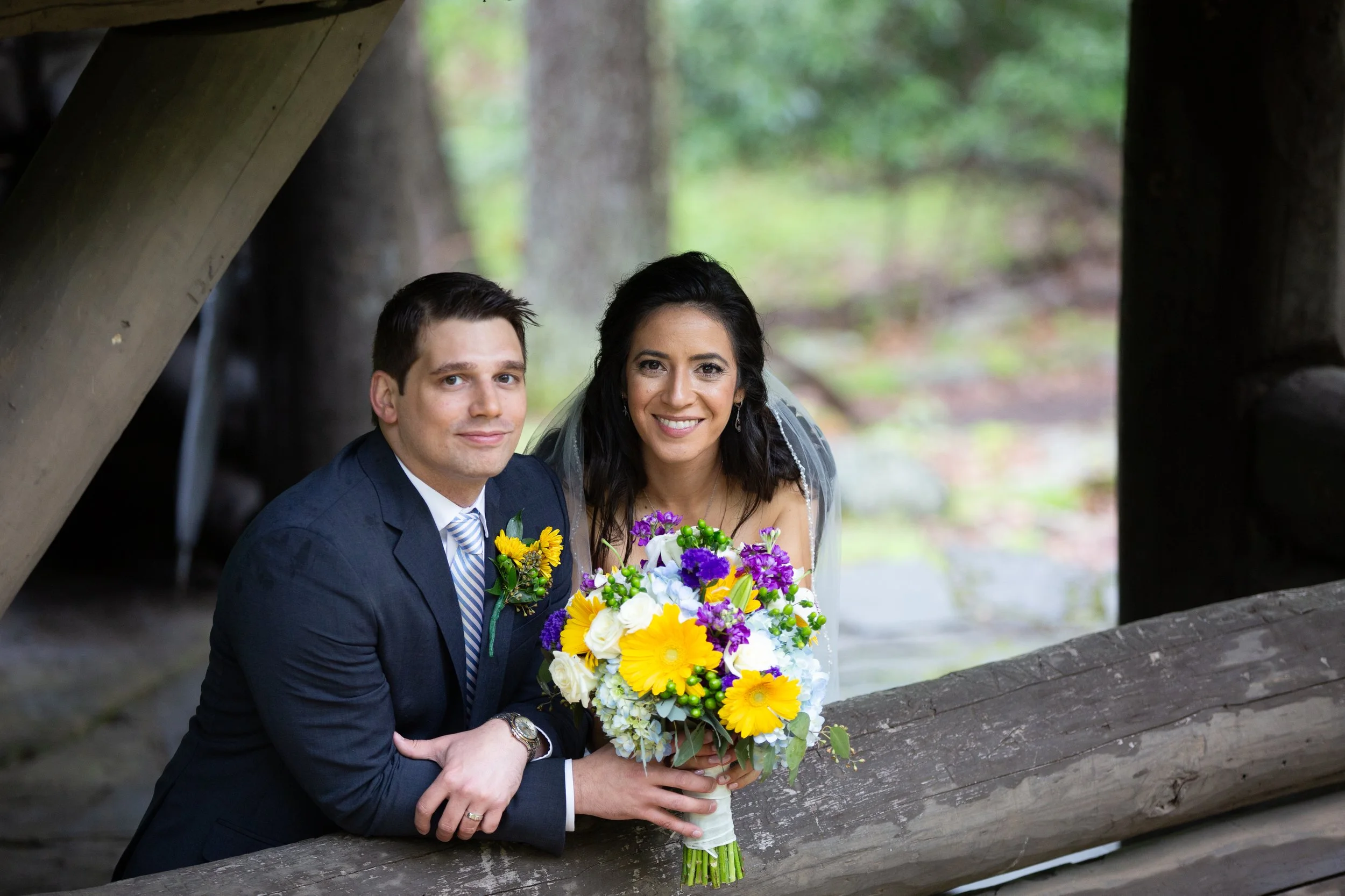 A couple dressed in wedding attire, holding a colorful bouquet, posing under a wooden bridge in a forested area.