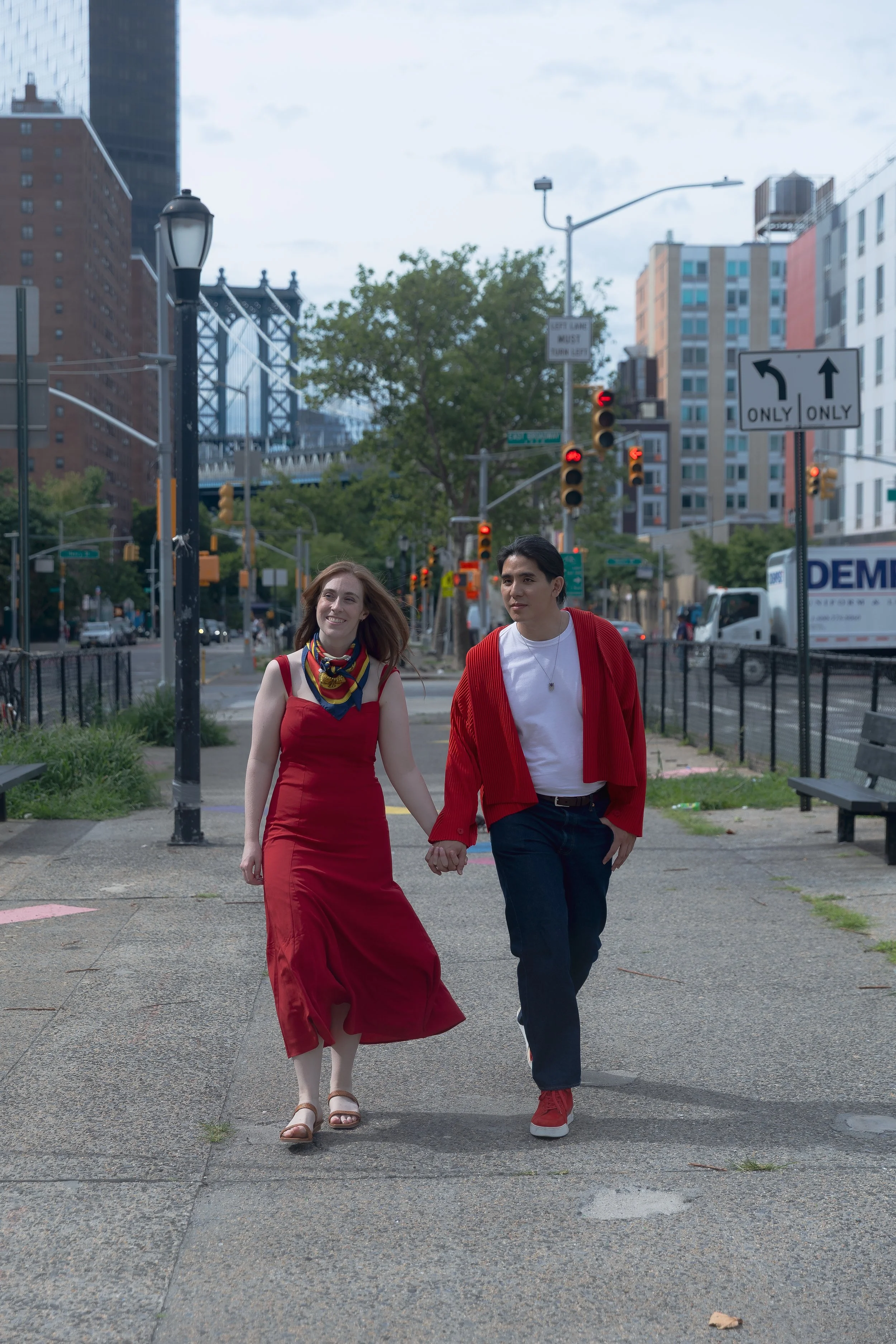 A woman in a red dress and a man in a red jacket and white shirt walk hand in hand on a city sidewalk during daytime, smiling and holding hands.
