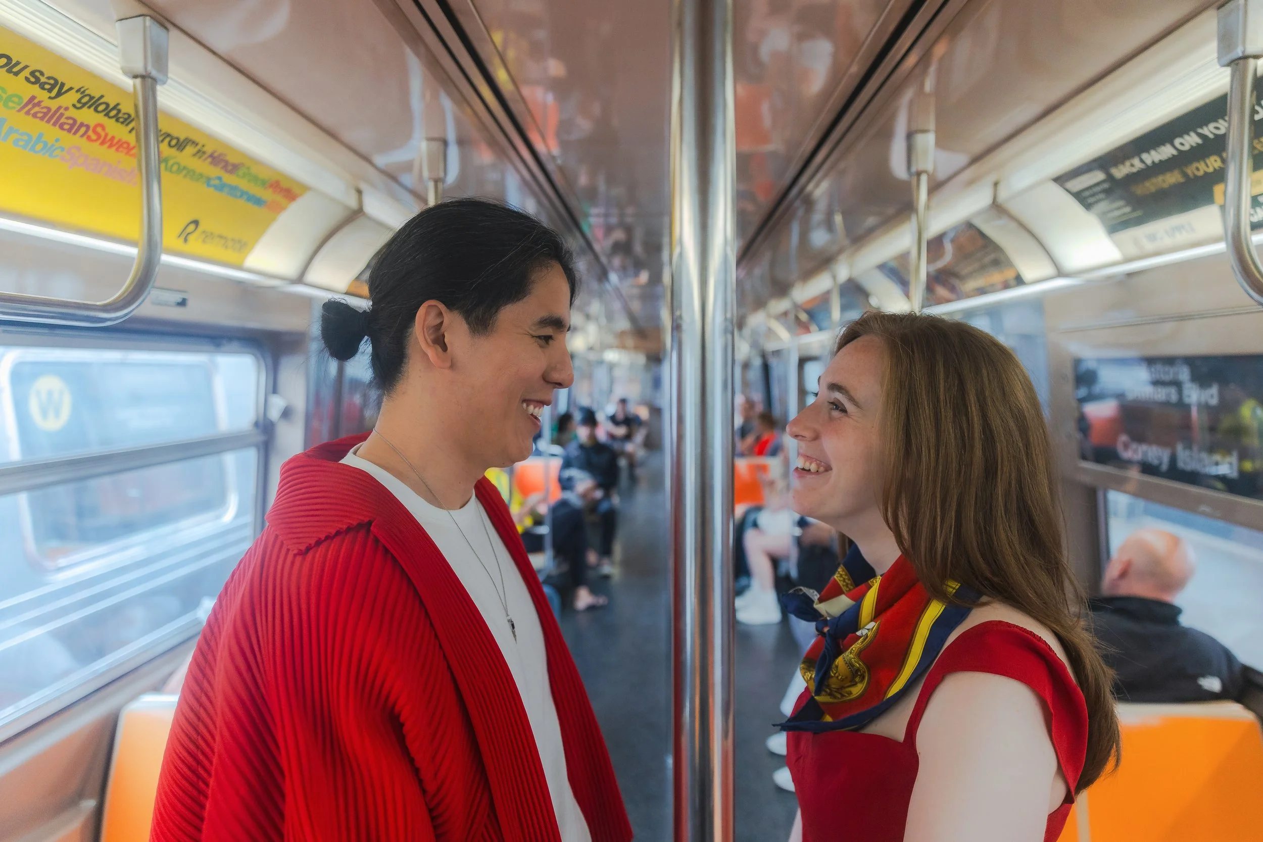 A couple in red outfits smiling at each other on a subway train, with other passengers in the background.