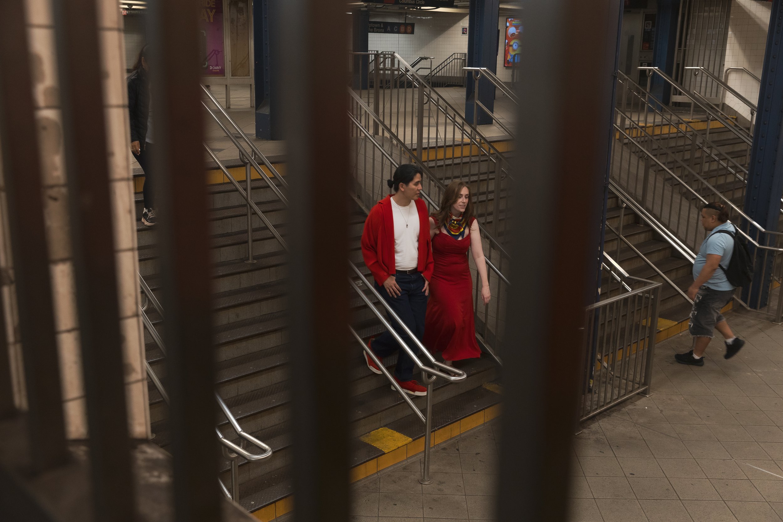 Two people walking down stairs in a subway station, one wearing a red dress and the other wearing a red jacket and white shirt, seen through metal bars.