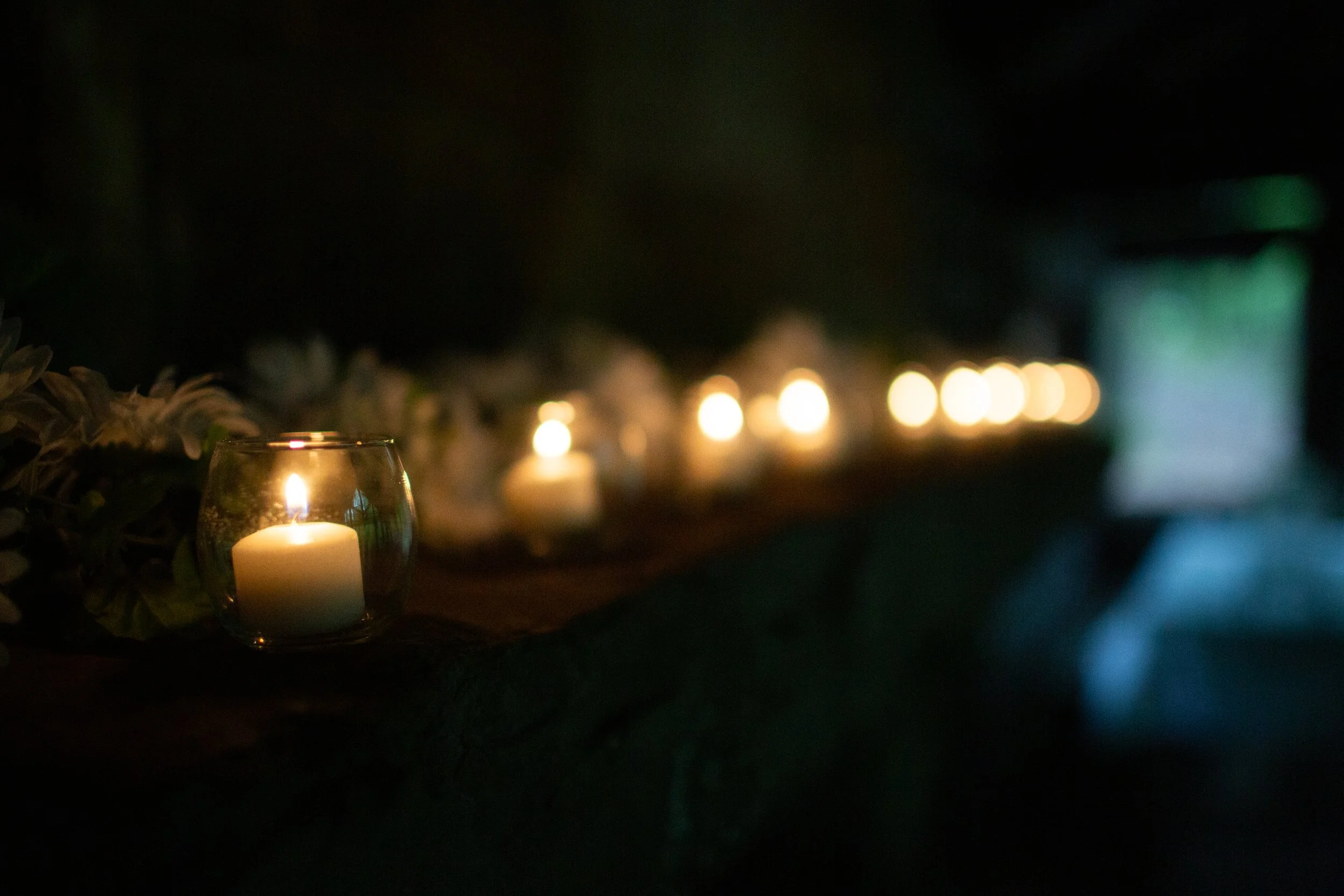 A candle inside a glass holder with a flower arrangement beside it, set on a dark surface with blurred candlelights in the background.