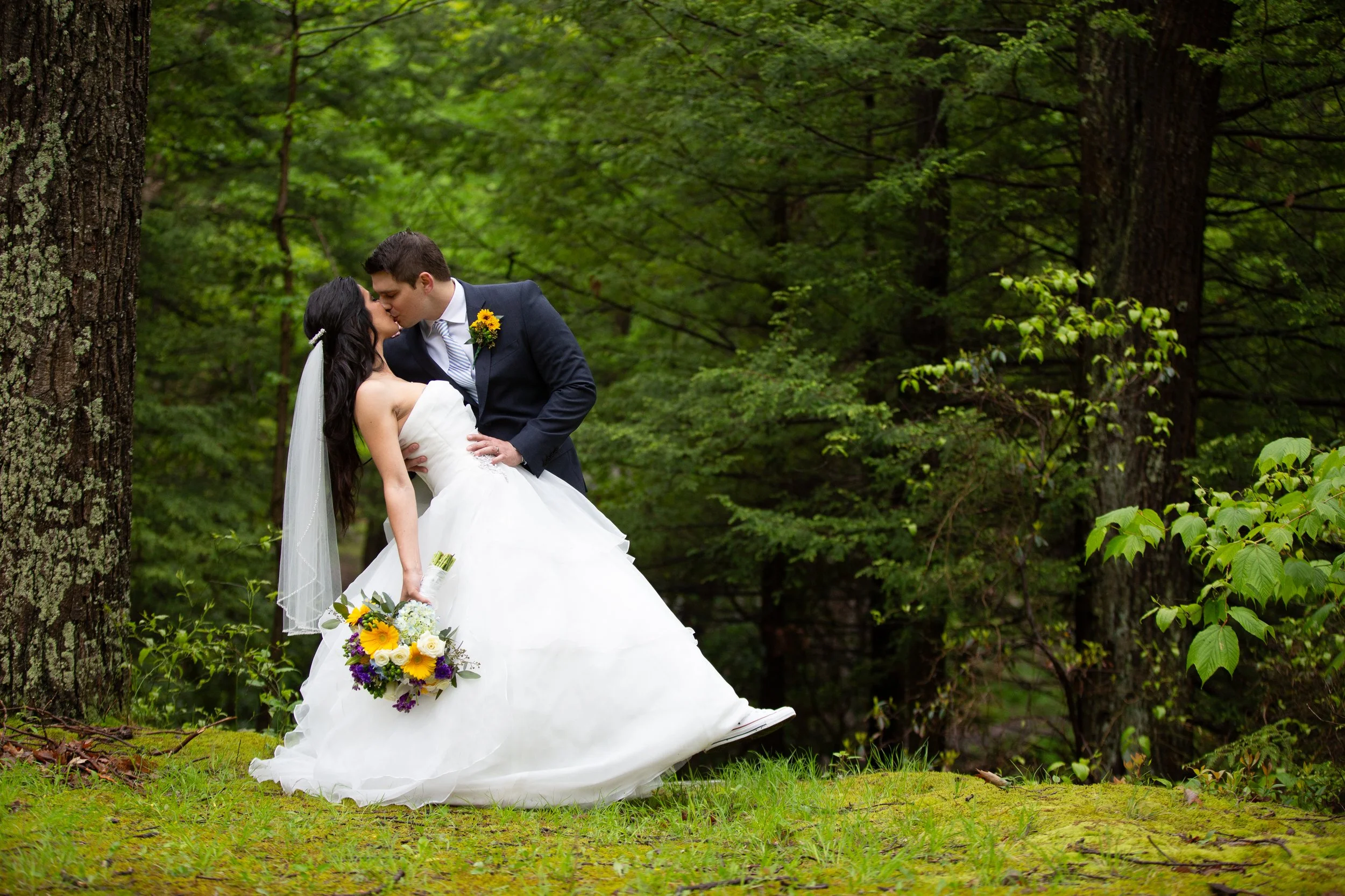 A bride and groom share a kiss in a wooded outdoor setting during their wedding, with the bride holding a bouquet of colorful flowers and the groom wearing a dark suit with a sunflower boutonniere.