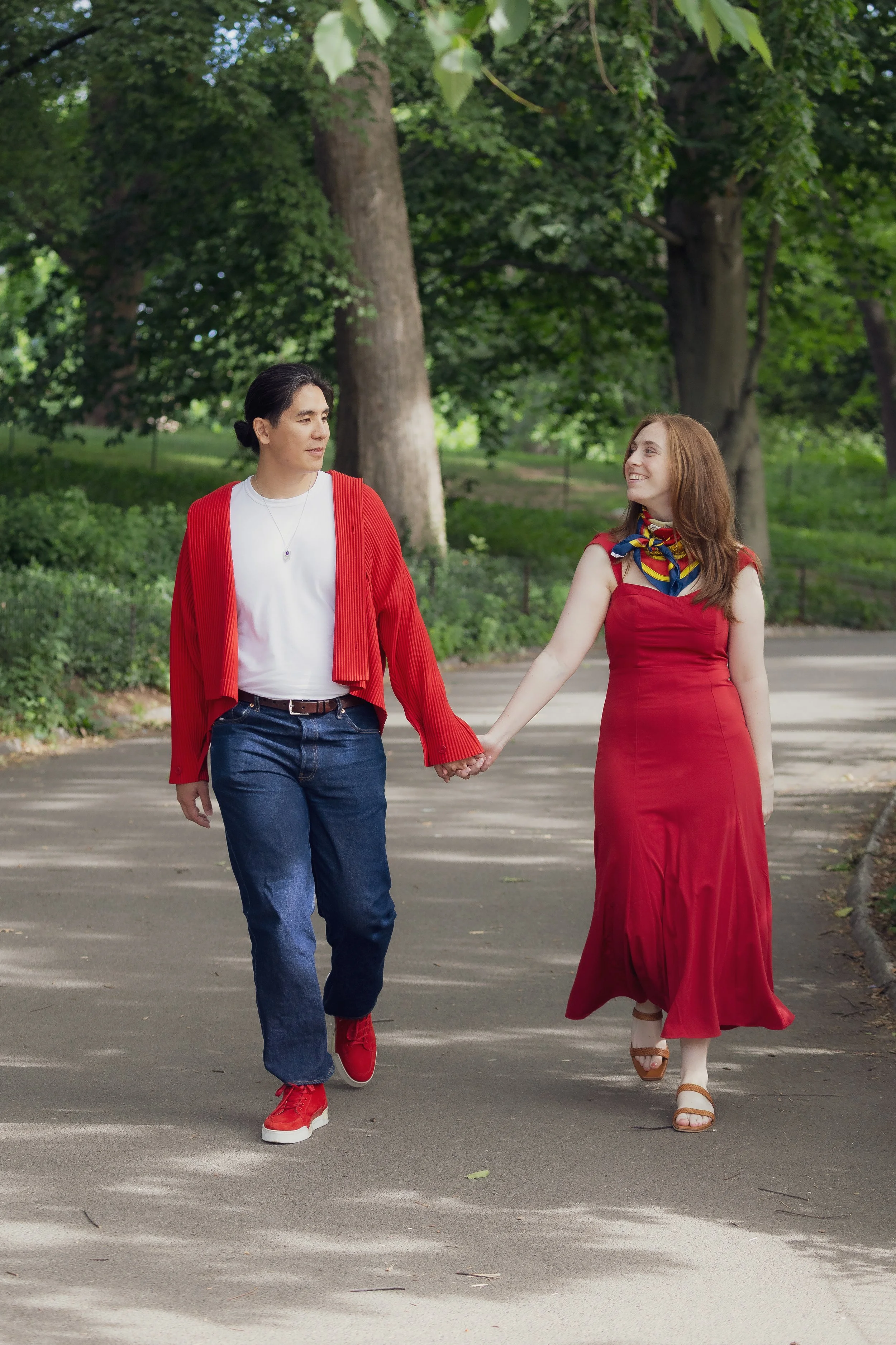 A man and woman holding hands while walking outdoors in a park with trees and greenery. The man is wearing a red cardigan, white t-shirt, and jeans, and the woman is wearing a red dress with a colorful scarf and white sandals.