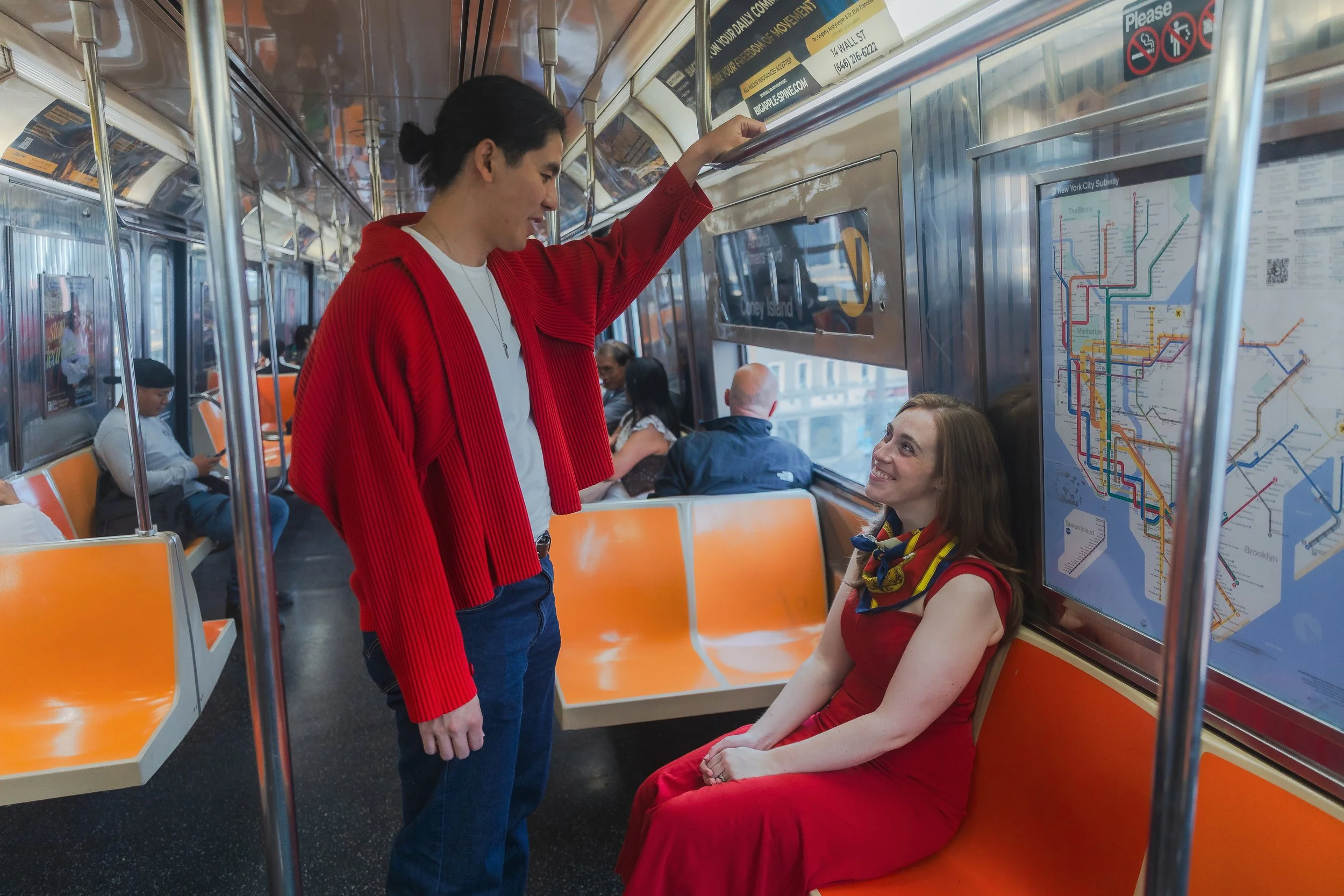 A young woman in a red dress listening to a man in a red sweater on a New York City subway train. The woman is seated, smiling, and the man is standing and leaning over her.
