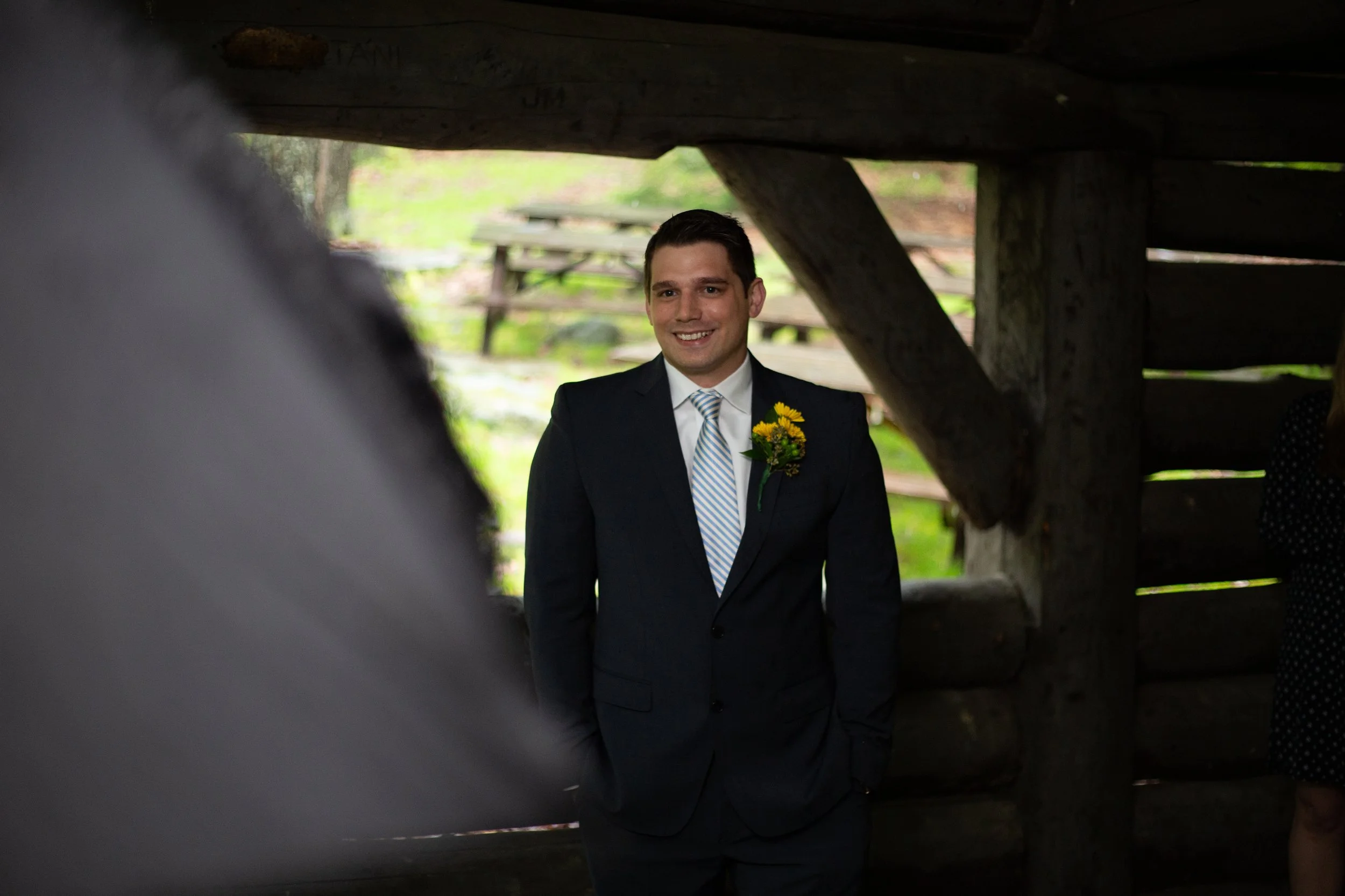 A man in a suit with a yellow flower boutonniere is smiling under a wooden structure, with picnic tables in the background in an outdoor setting.