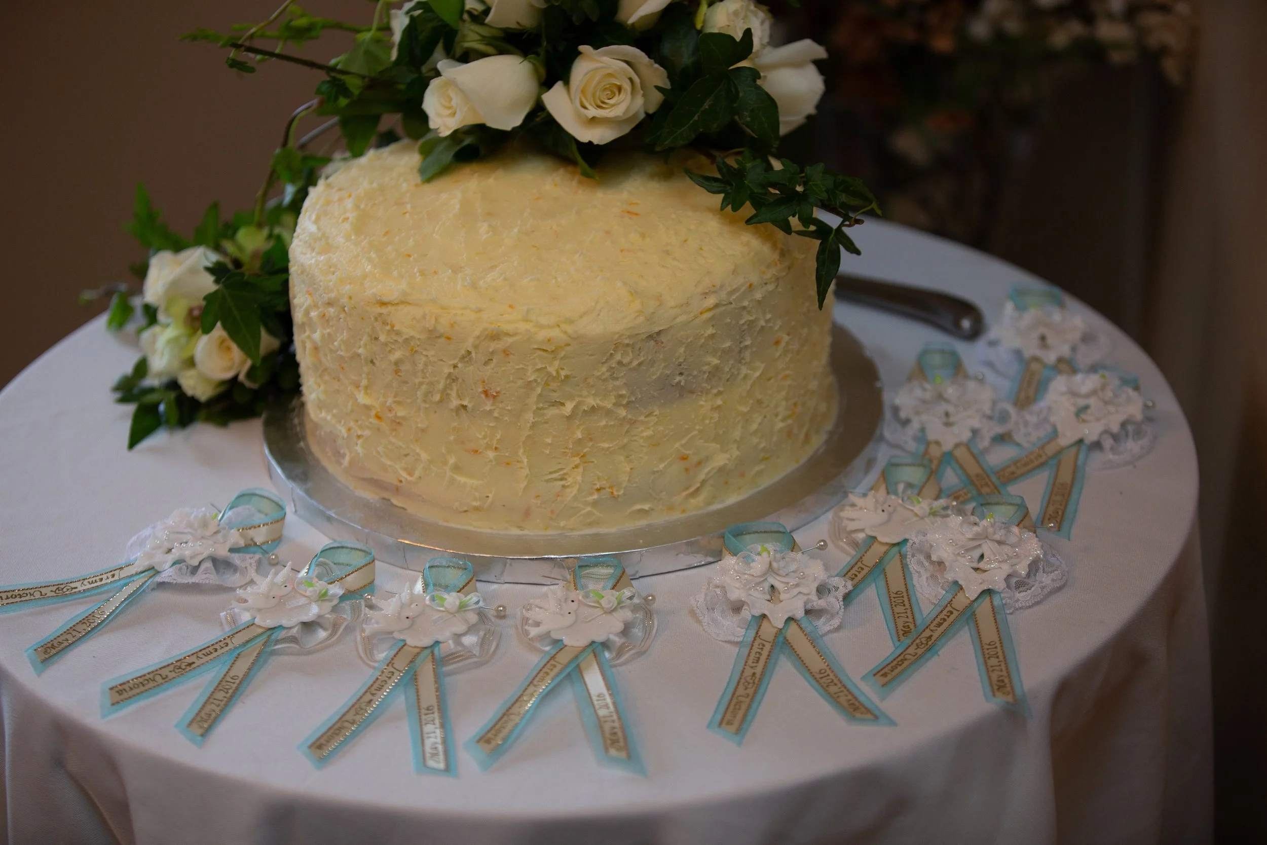 A wedding cake with white roses and green foliage on top, decorated with light blue and gold ribbons and small edible flowers, is placed on a table with a white tablecloth surrounded by matching wedding favors.