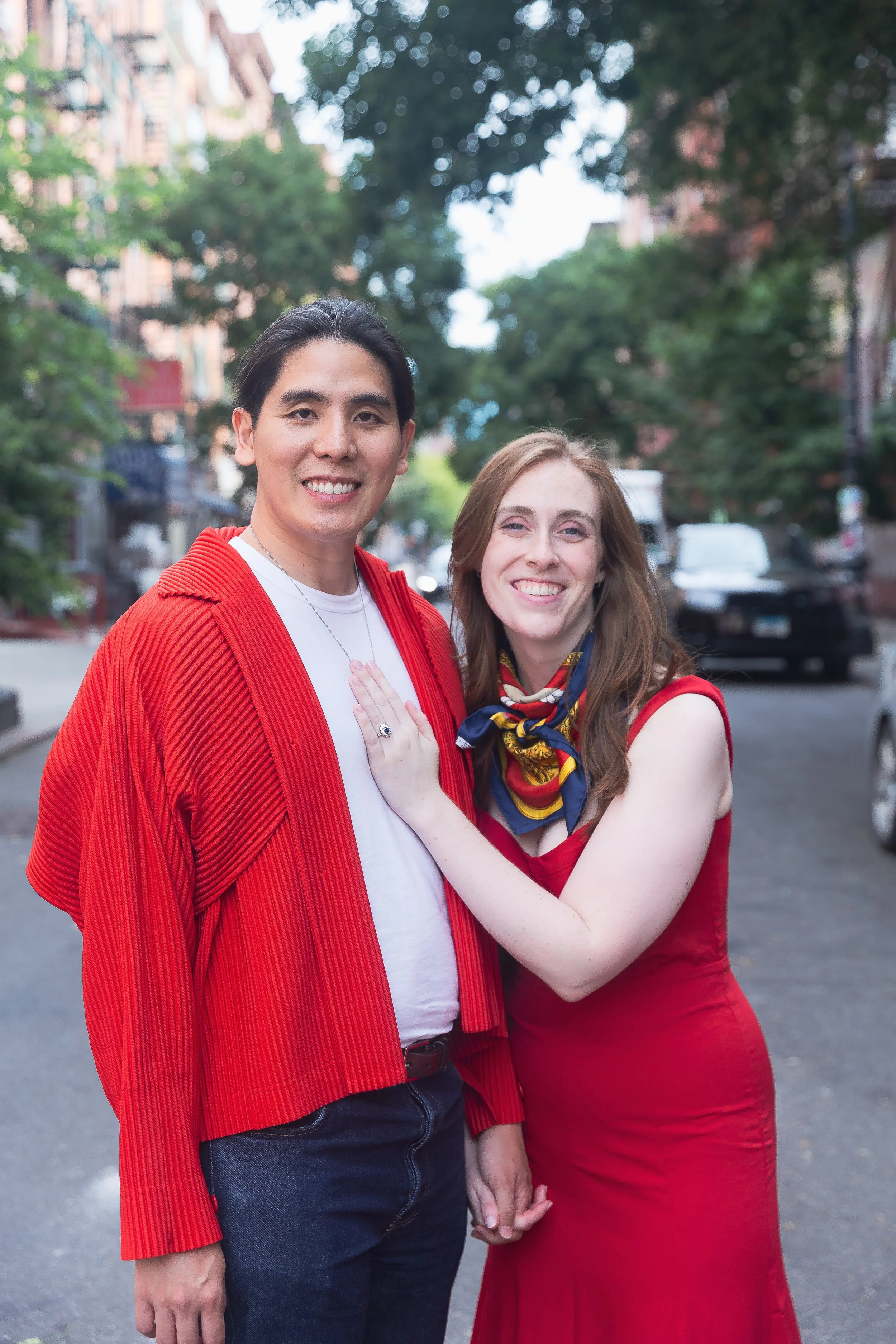A man and woman standing outdoors on a street, holding hands, smiling at the camera, with trees and cars in the background, both dressed in red.