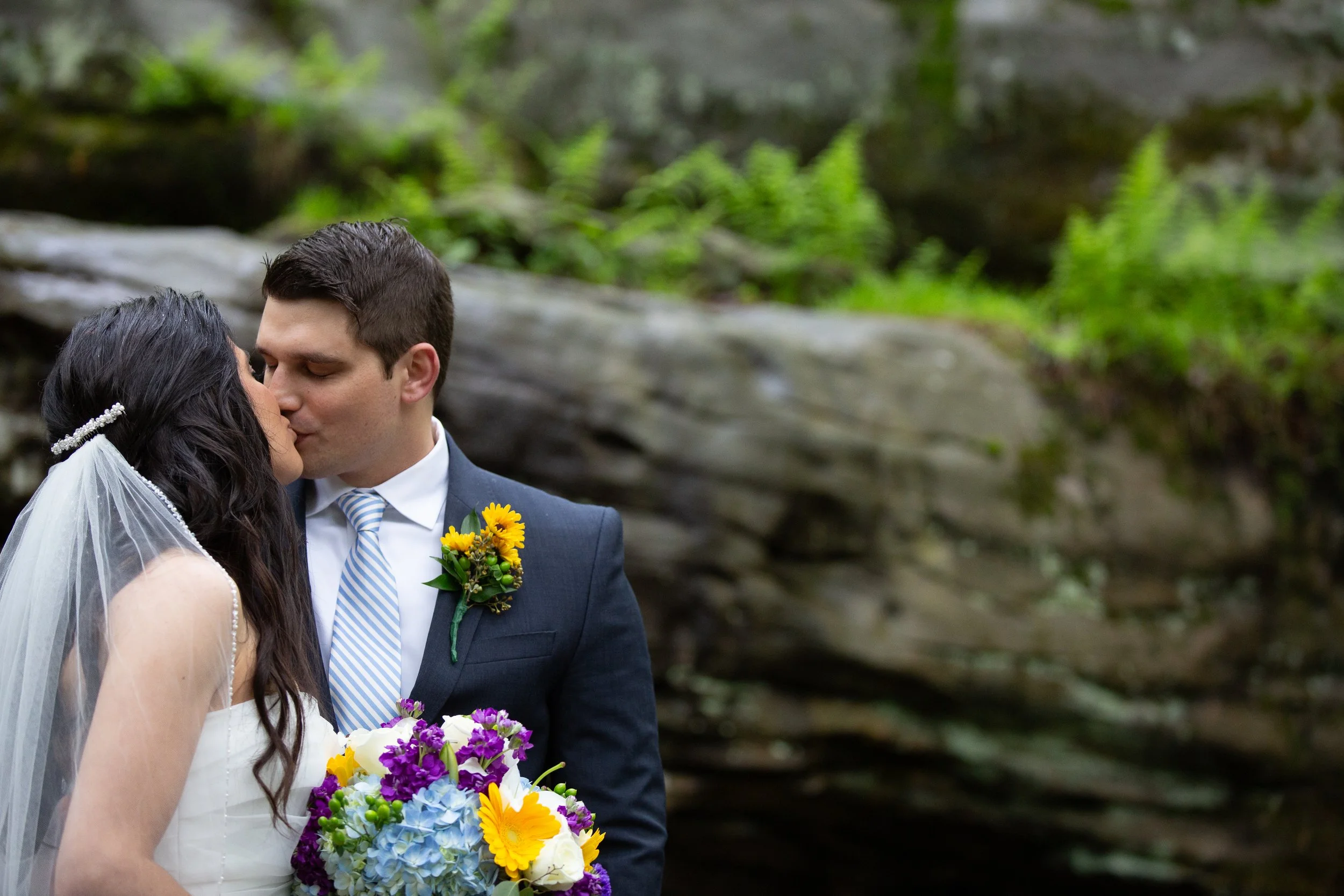 A bride and groom share a kiss outdoors surrounded by green foliage, with the bride holding a colorful bouquet of flowers.