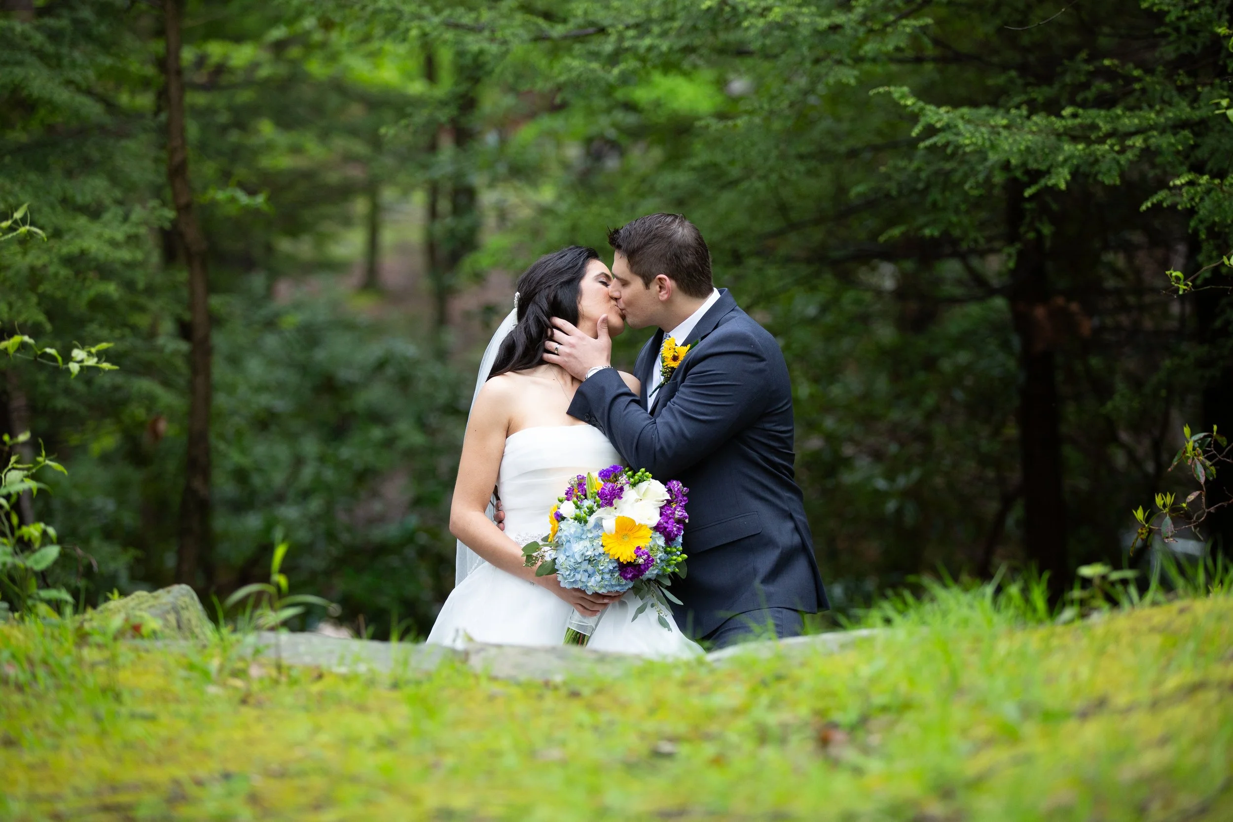 A bride and groom kissing in a lush, green outdoor setting during their wedding, with the bride holding a colorful bouquet of flowers.