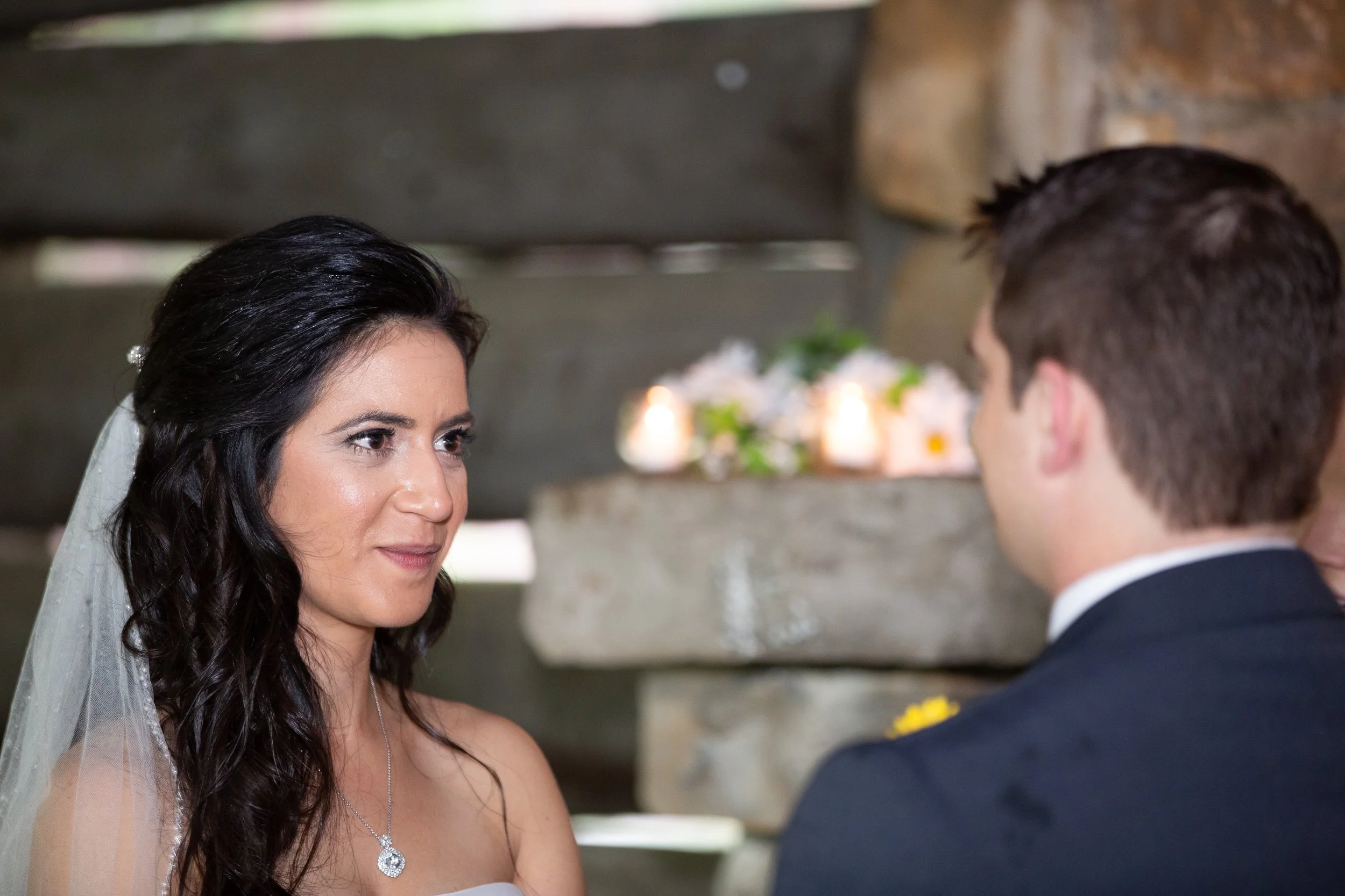 A bride with dark, wavy hair and a veil gazes at a groom in a tuxedo during a wedding ceremony.