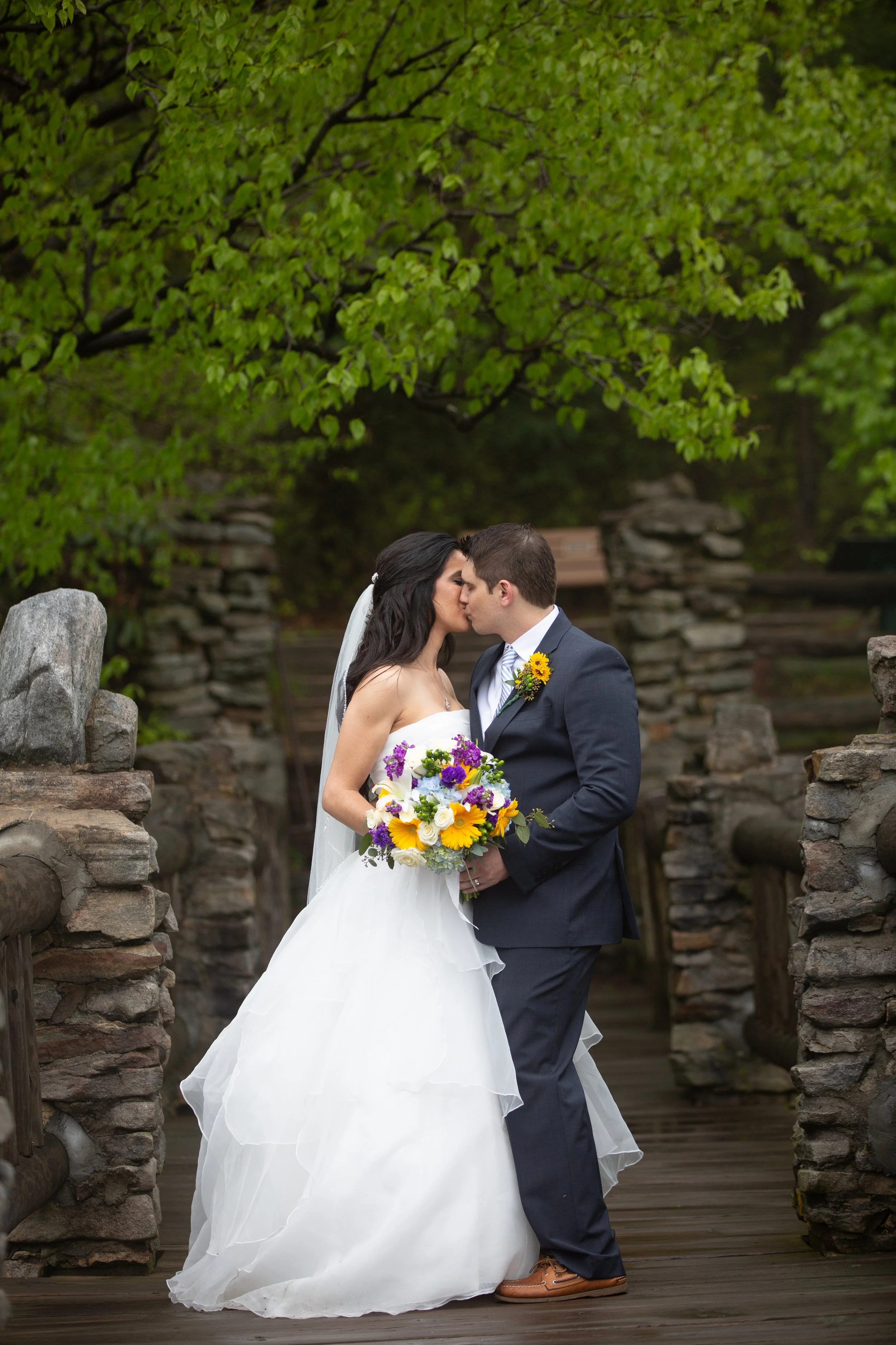 A bride and groom share a kiss outdoors on a stone bridge beneath a leafy green tree, with the bride holding a colorful bouquet and the groom wearing a suit with a sunflower boutonniere.