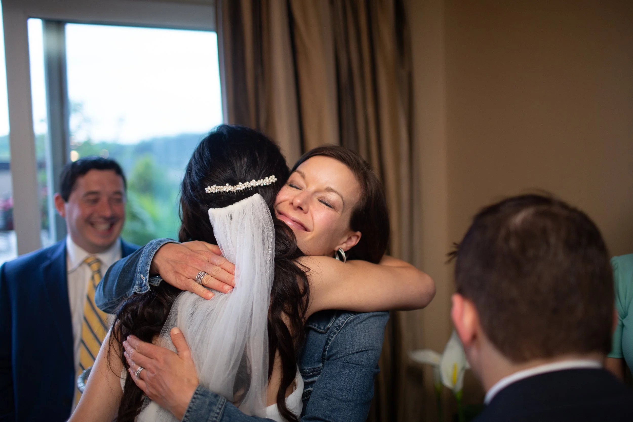 Two women hugging, one with long dark hair and a veil, the other with short brown hair, in a room with large windows and gold curtains, a man in a suit smiling in the background, and a person with short hair in the foreground.