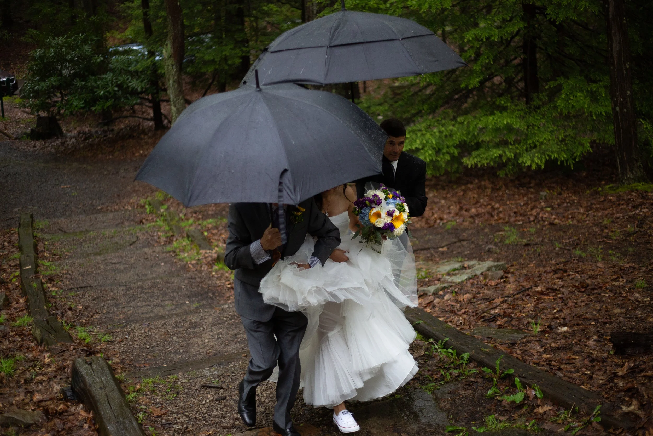 A bride in a wedding dress with a bouquet of flowers and a groom in a suit walking outside in a wooded area, holding umbrellas in the rain.