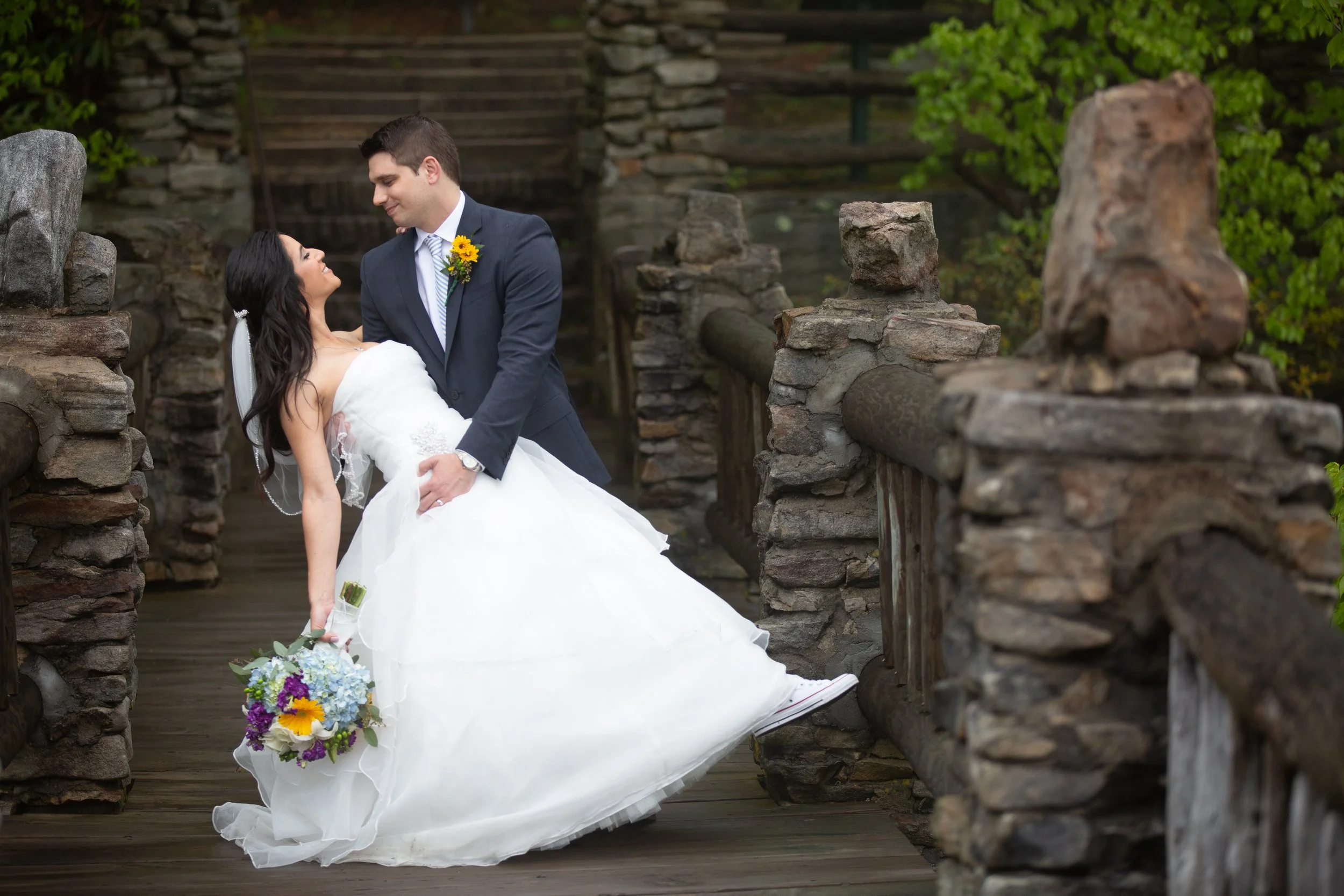 A bride and groom are dancing on a rustic stone bridge, with the bride holding a colorful bouquet and the groom wearing a suit with a sunflower boutonniere.