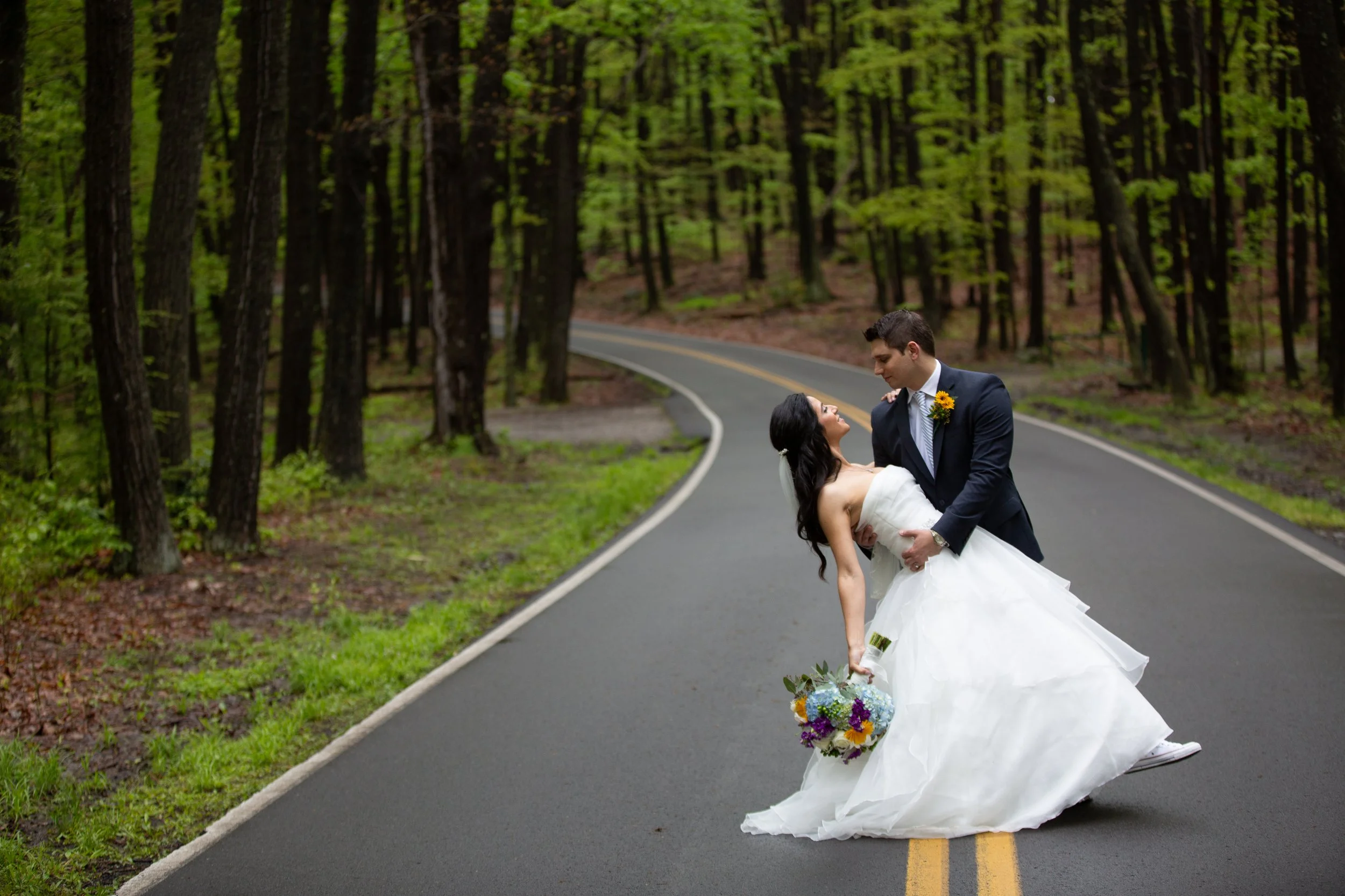 A bride and groom are on a winding forest road, with the groom dipping the bride as she holds a bouquet of flowers, in a romantic moment during their wedding photoshoot.
