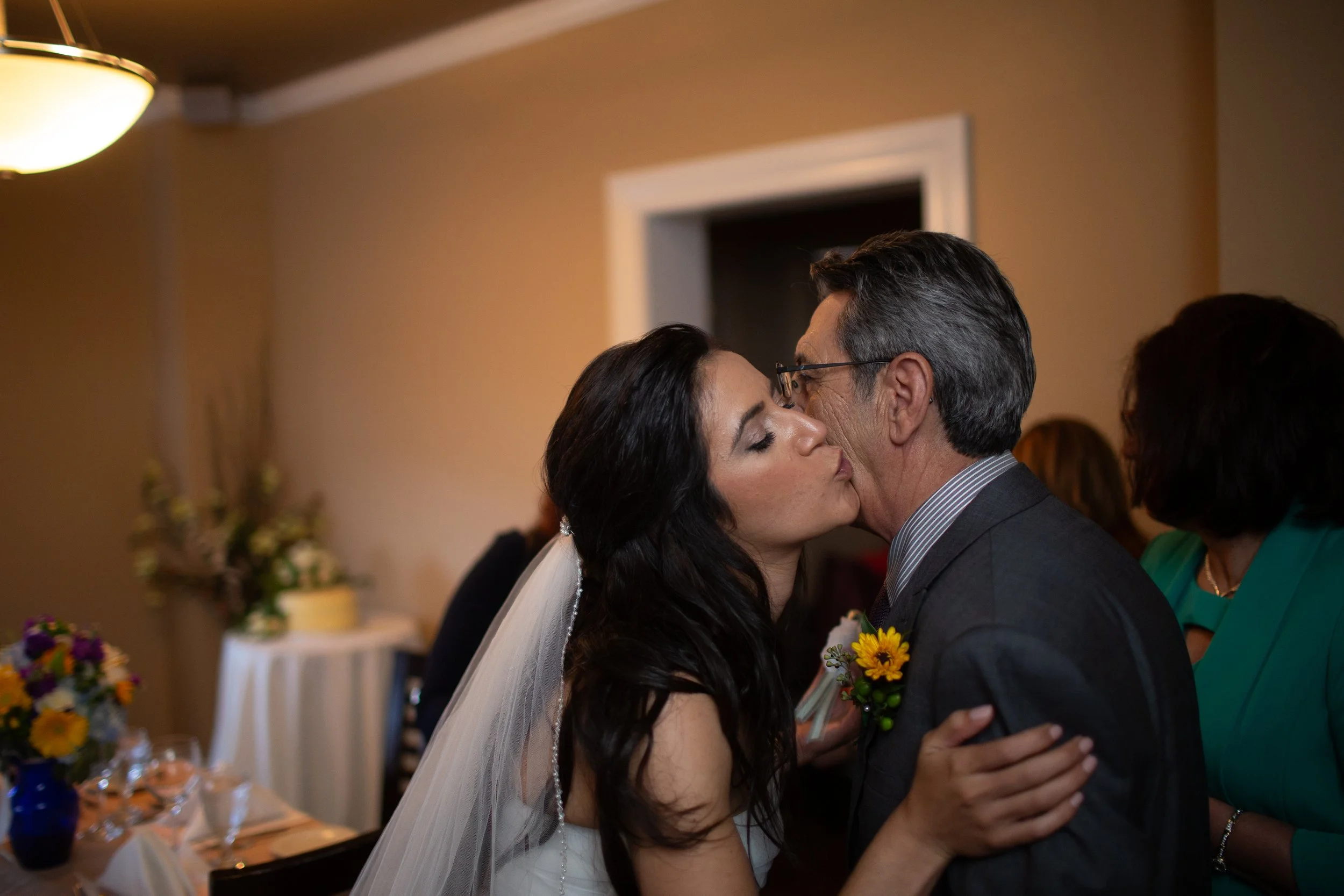 A bride and groom sharing a kiss during their wedding reception. The bride has dark hair and a veil; the groom has gray hair and glasses. Guests are in the background, and floral arrangements are on tables.