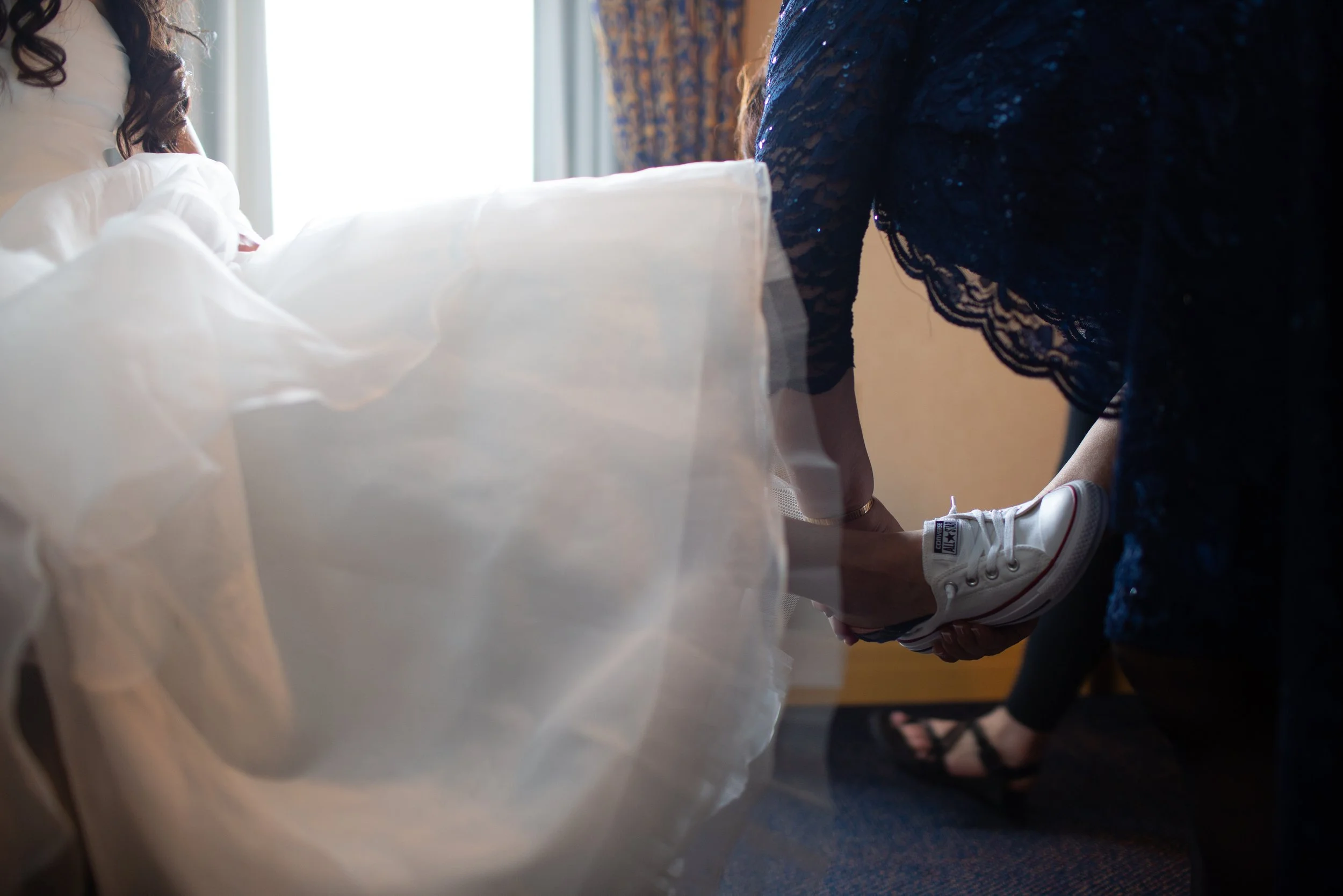 A person wearing a dark lace dress and white sneakers, kneeling on the floor, holding hands with another person whose arm is visible, in front of a bed with white bedding near a window with sunlight.