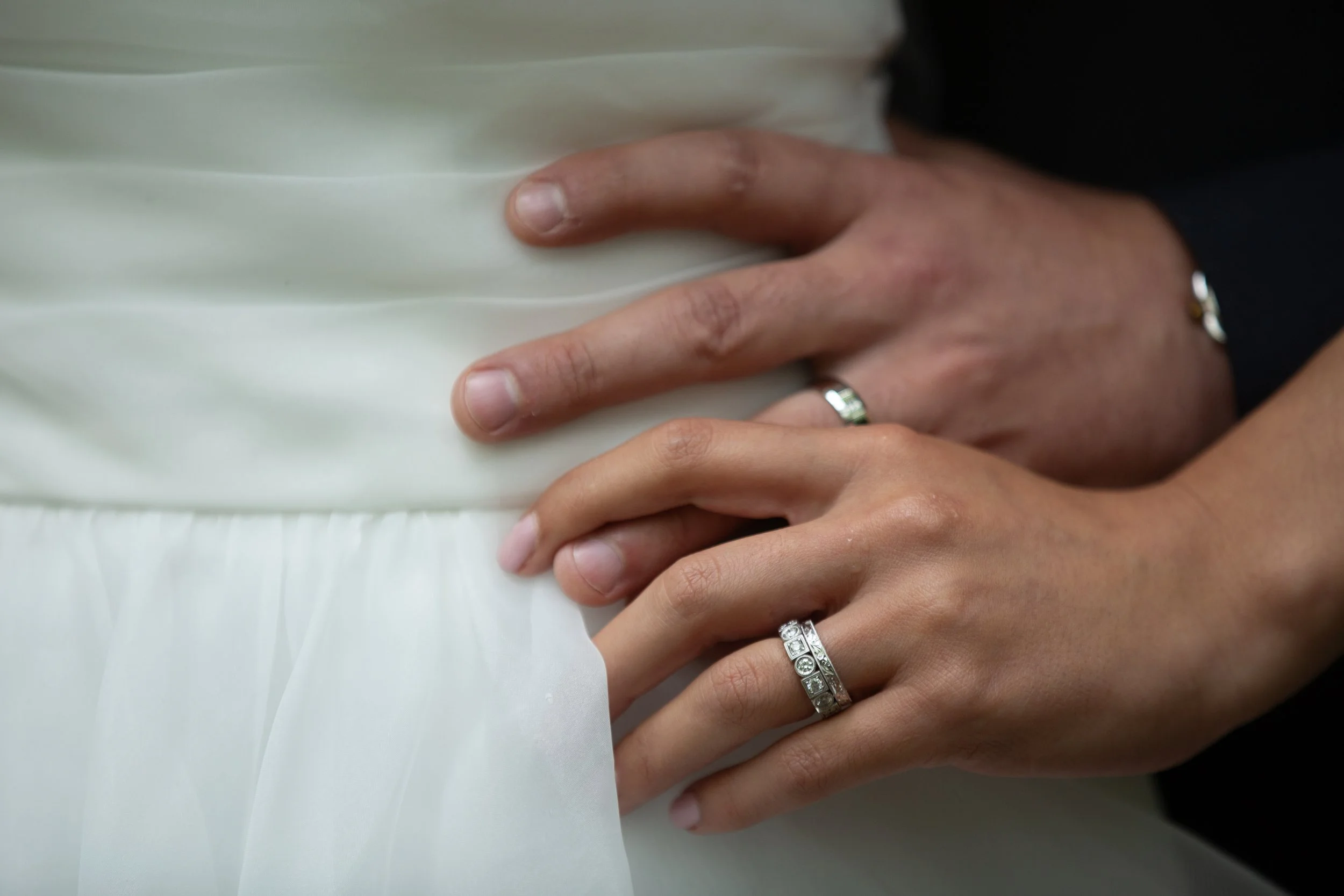 Close-up of a couple's hands, with rings, gently placed on the bride's white gown.