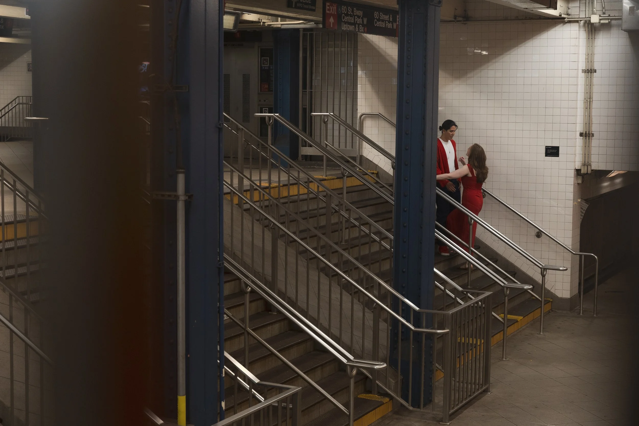 Subway entrance with stairs and handrails, two women wearing red dresses standing and talking near the railing in an underground station with tiled walls and directional signs overhead.