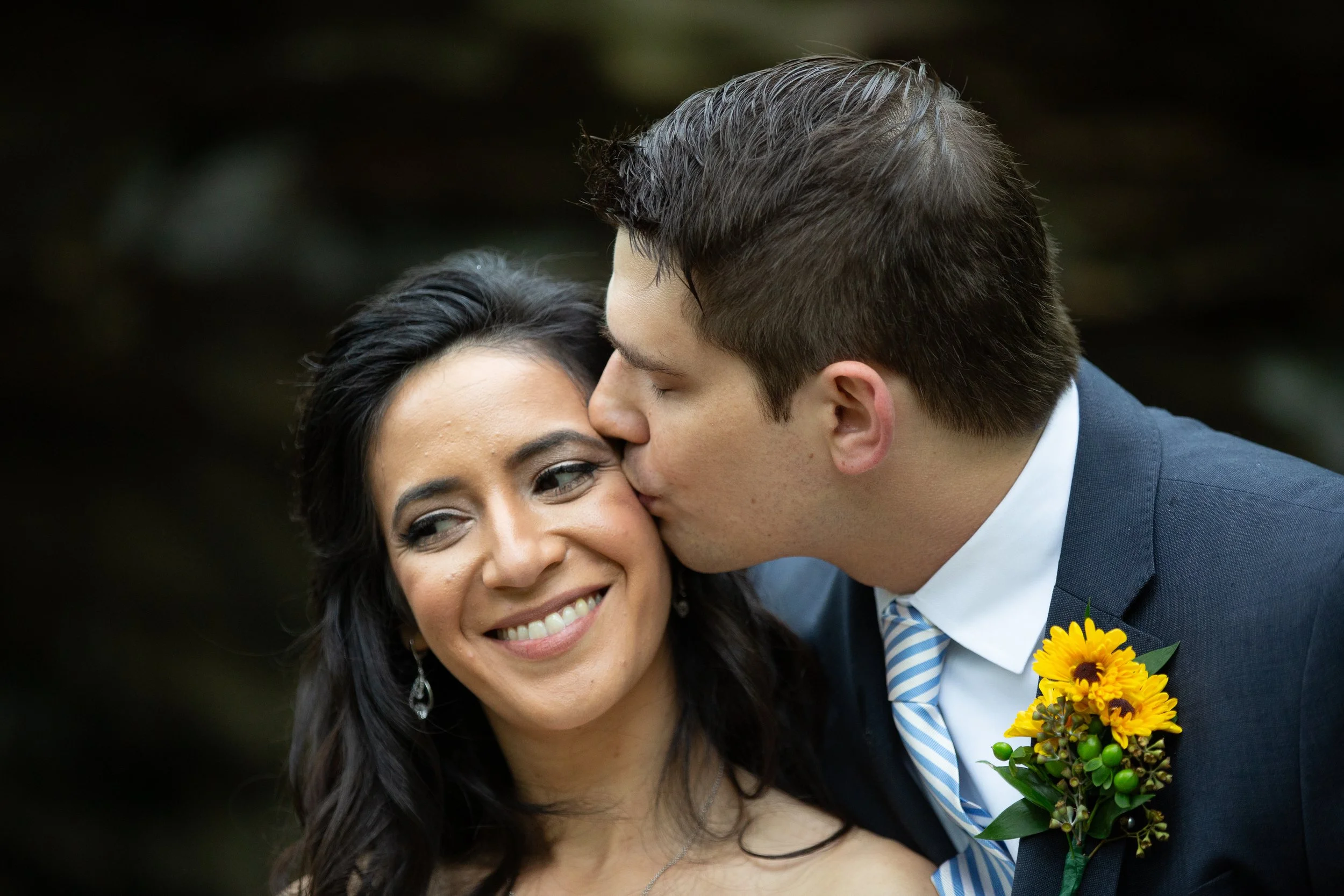 A man in a suit kisses a woman on the cheek during a celebration, holding a yellow sunflower bouquet.