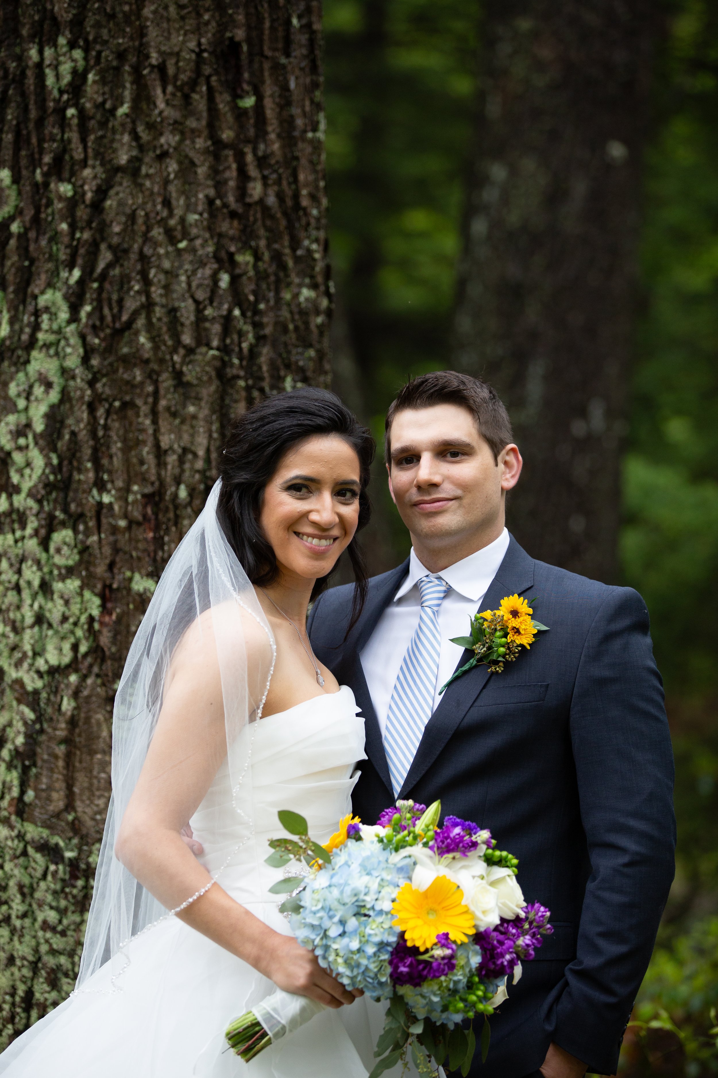 A bride and groom standing outdoors in front of a large tree, smiling at the camera. The bride is wearing a white wedding dress and veil, holding a colorful bouquet of flowers. The groom is wearing a dark suit with a light-colored tie and a sunflower