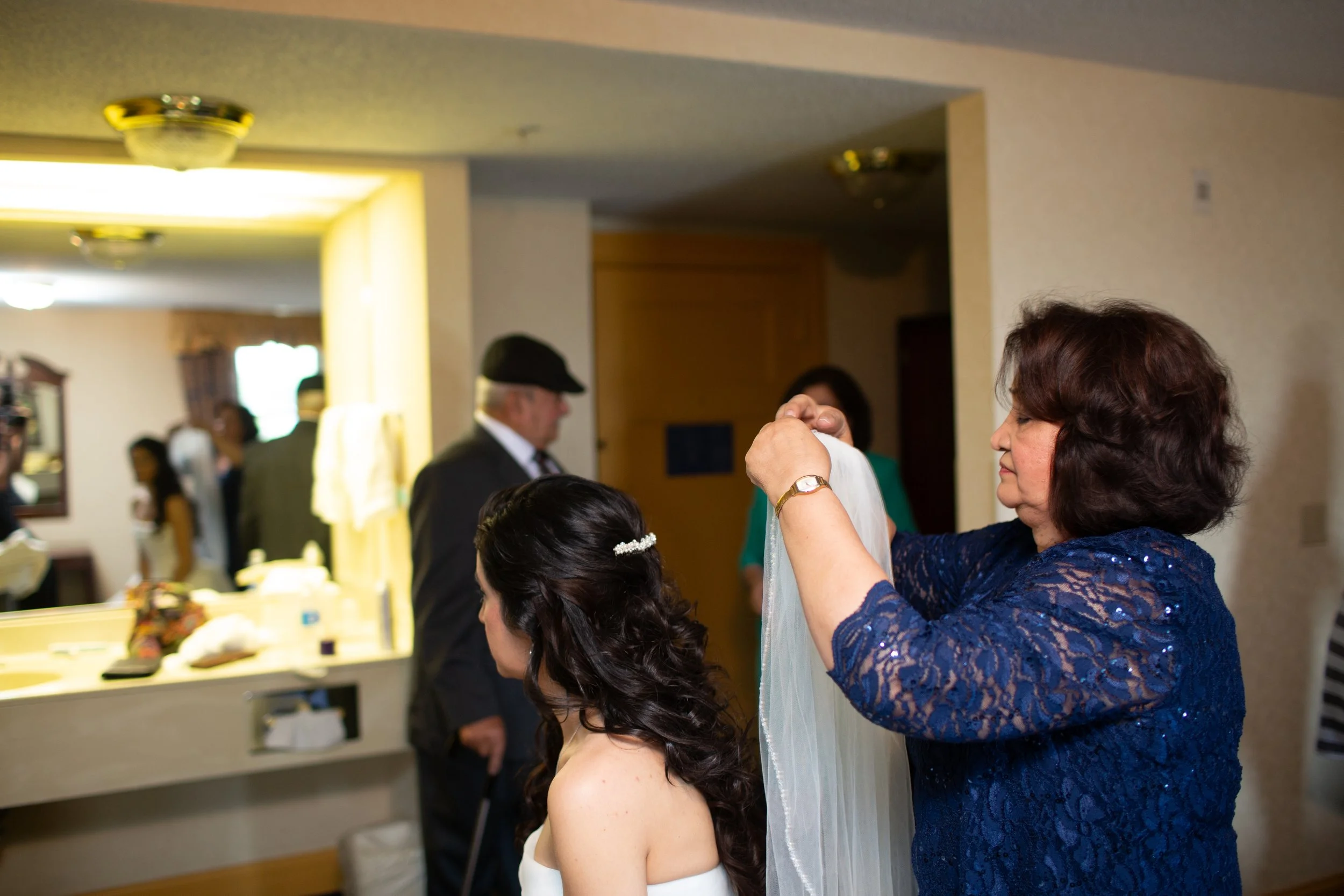 Women preparing wedding dress on bride during wedding preparation in a room with mirror and people in background.