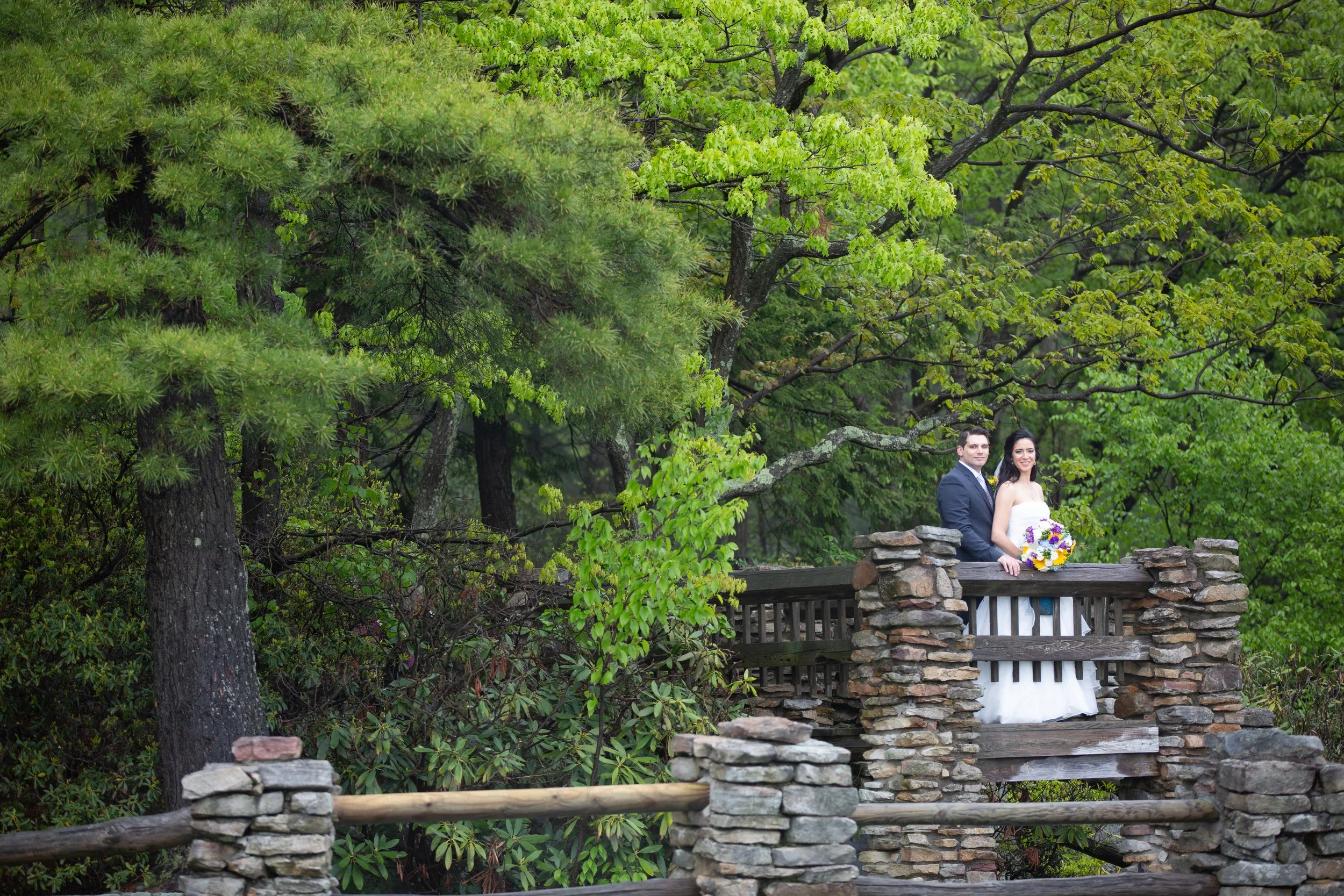 A bride and groom standing on a stone and wooden bridge surrounded by lush green trees and foliage.