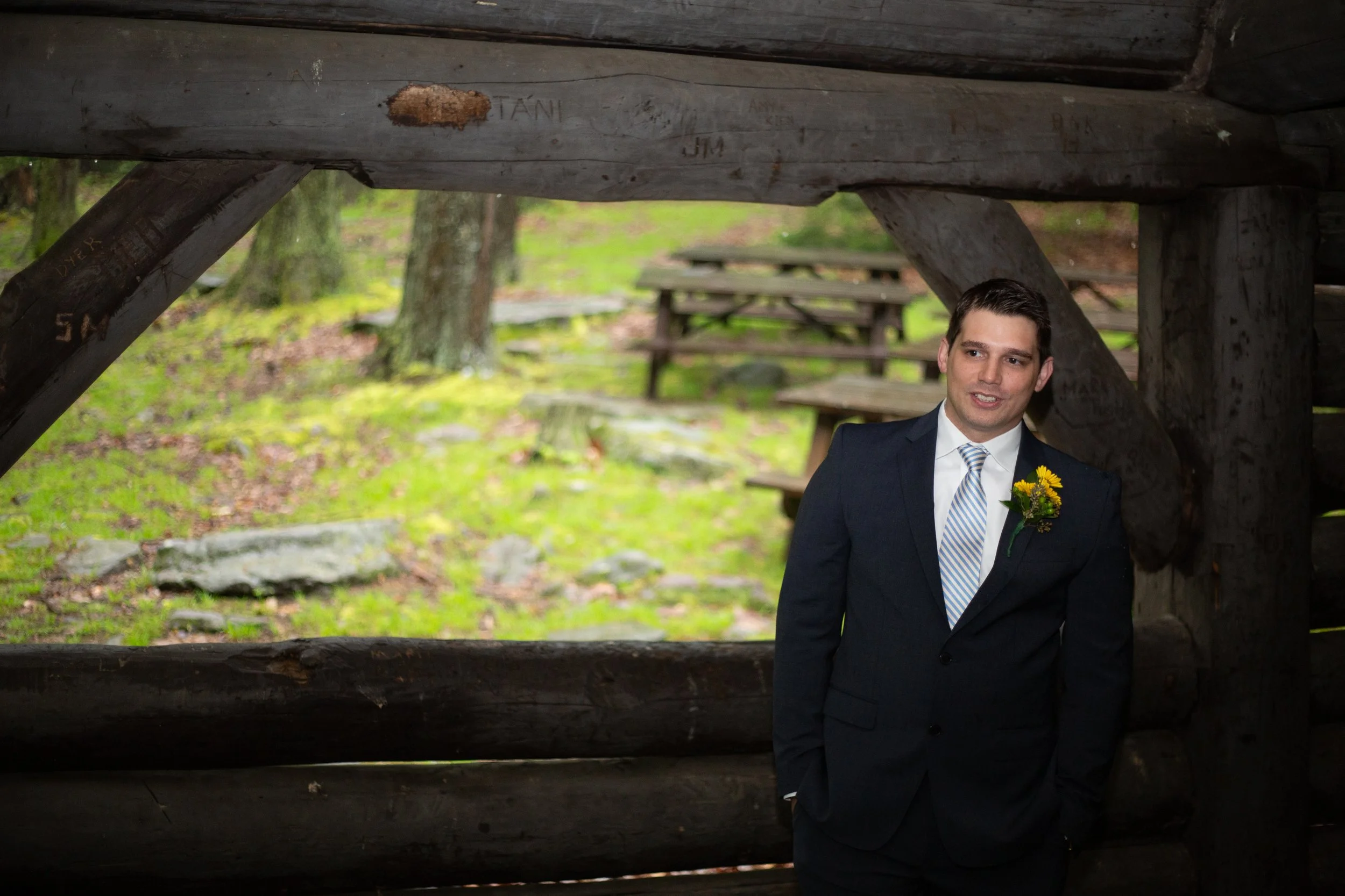 A man in a dark suit and light-colored striped tie with a small yellow flower boutonniere stands outside under a rustic wooden shelter with additional picnic tables in the background.
