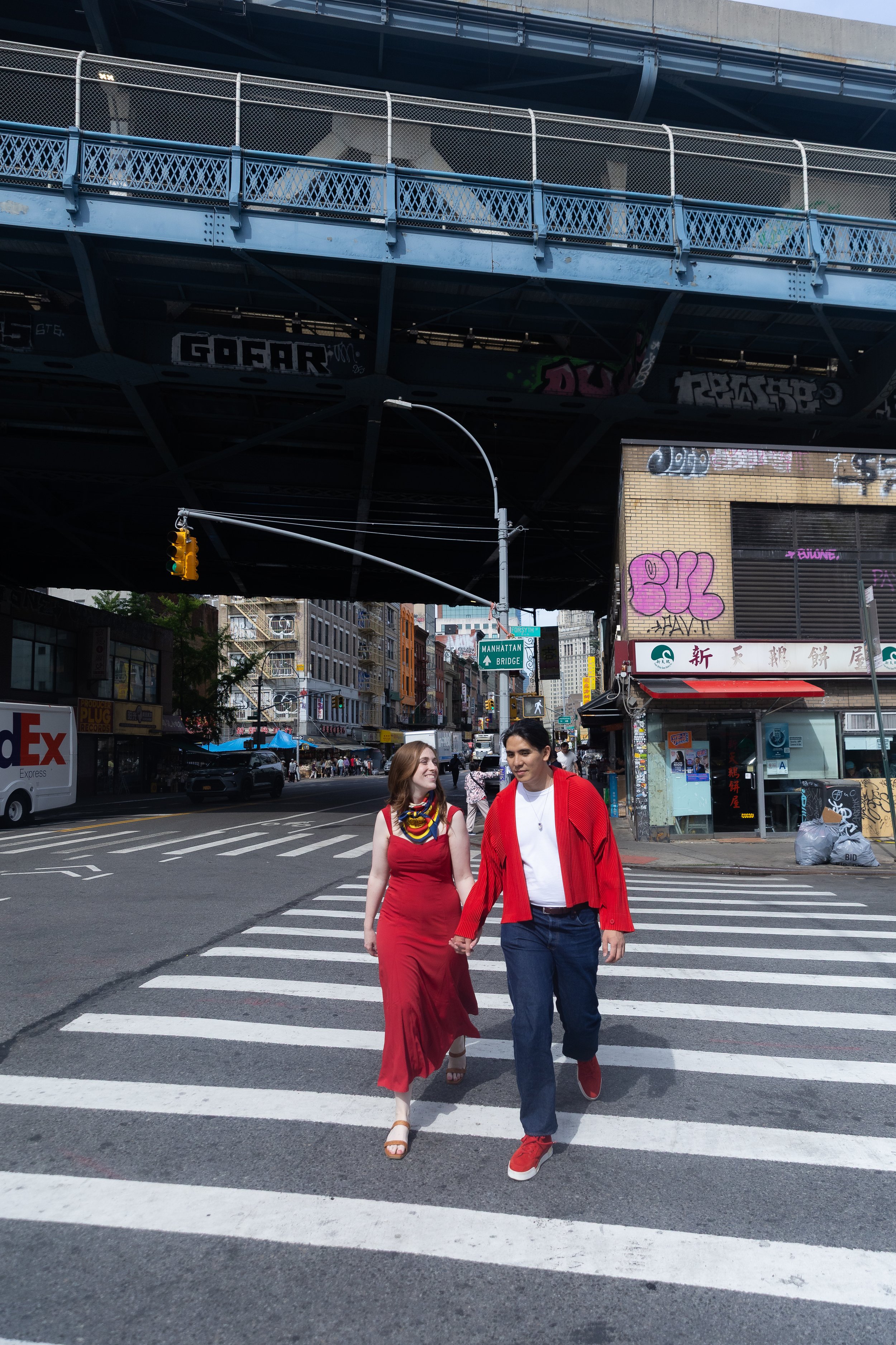 A young couple holding hands and walking across a city crosswalk, with a highway overpass above them and buildings and street signs in the background. The woman is wearing a red dress and beige sandals, and the man is wearing a red jacket, white shir