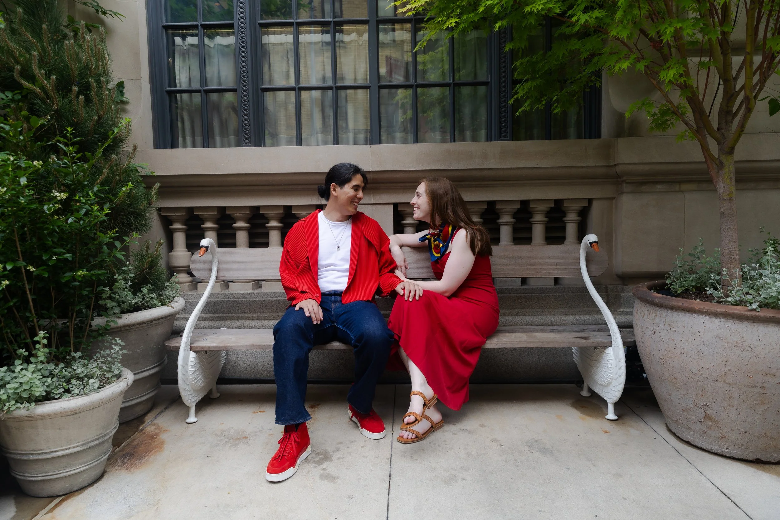 Two women sitting on a bench outdoors, smiling and looking at each other. One woman wears a red sweater and jeans, the other wears a red dress and sandals. Plants and trees surround the bench.