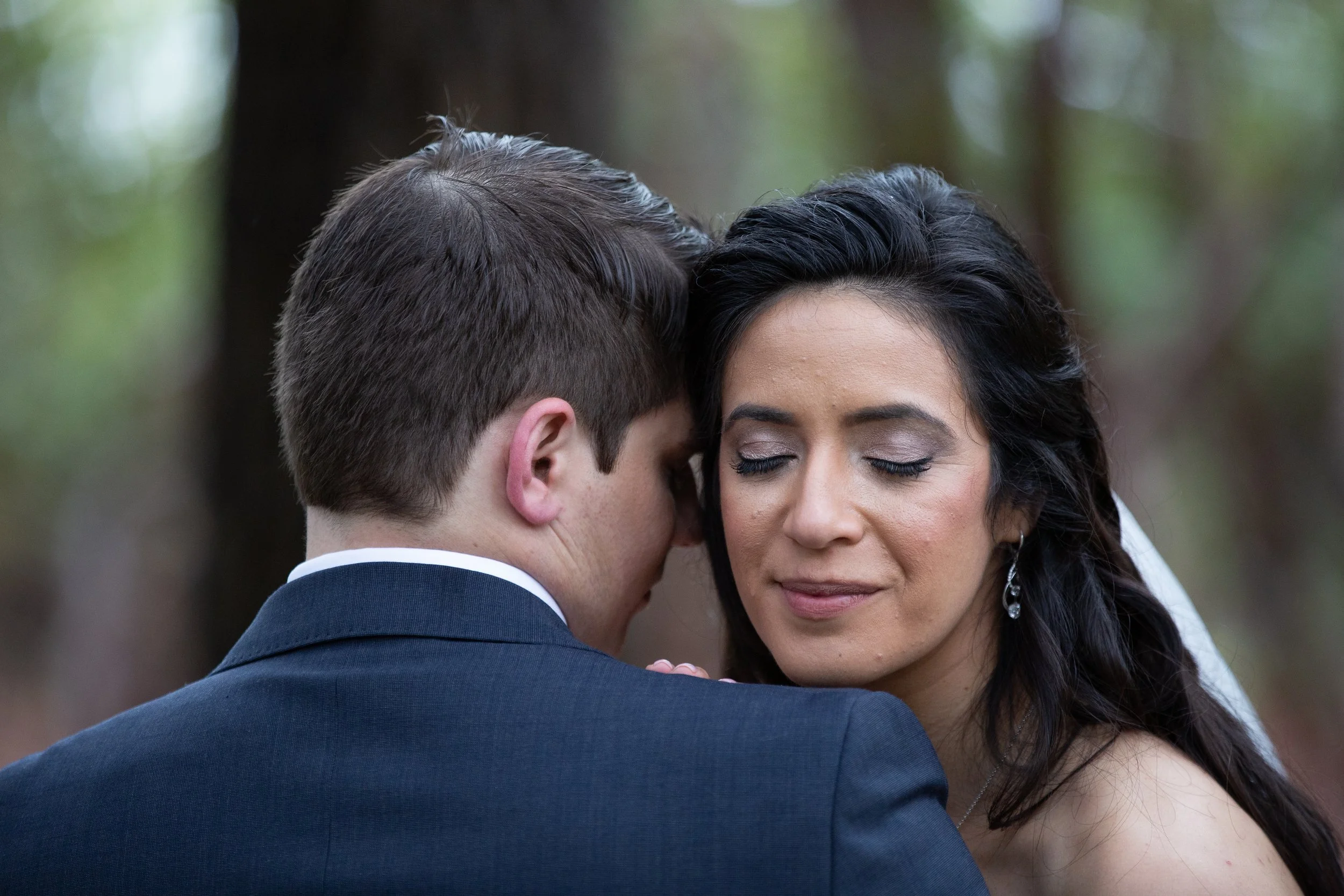 A close-up of a couple during a wedding ceremony, with the groom whispering into the bride's ear, both with their eyes closed, in an outdoor setting.