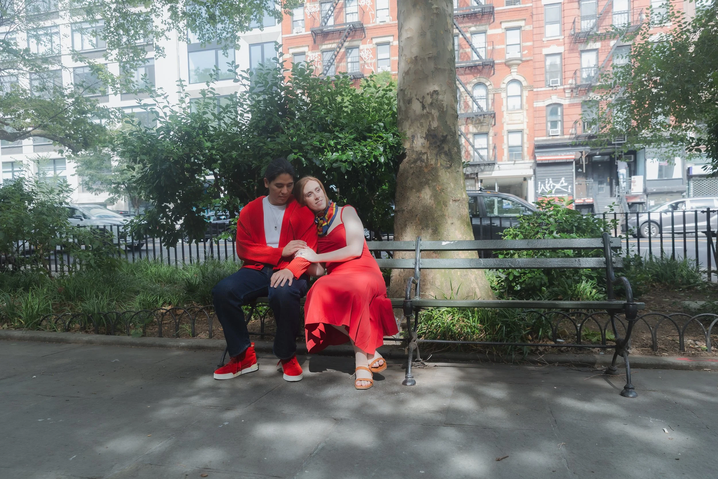 Two people sitting on a park bench; one wearing a red coat and dark pants, the other in a red dress with a colorful scarf, resting their heads together. They are in an urban park with trees, greenery, and buildings in the background.