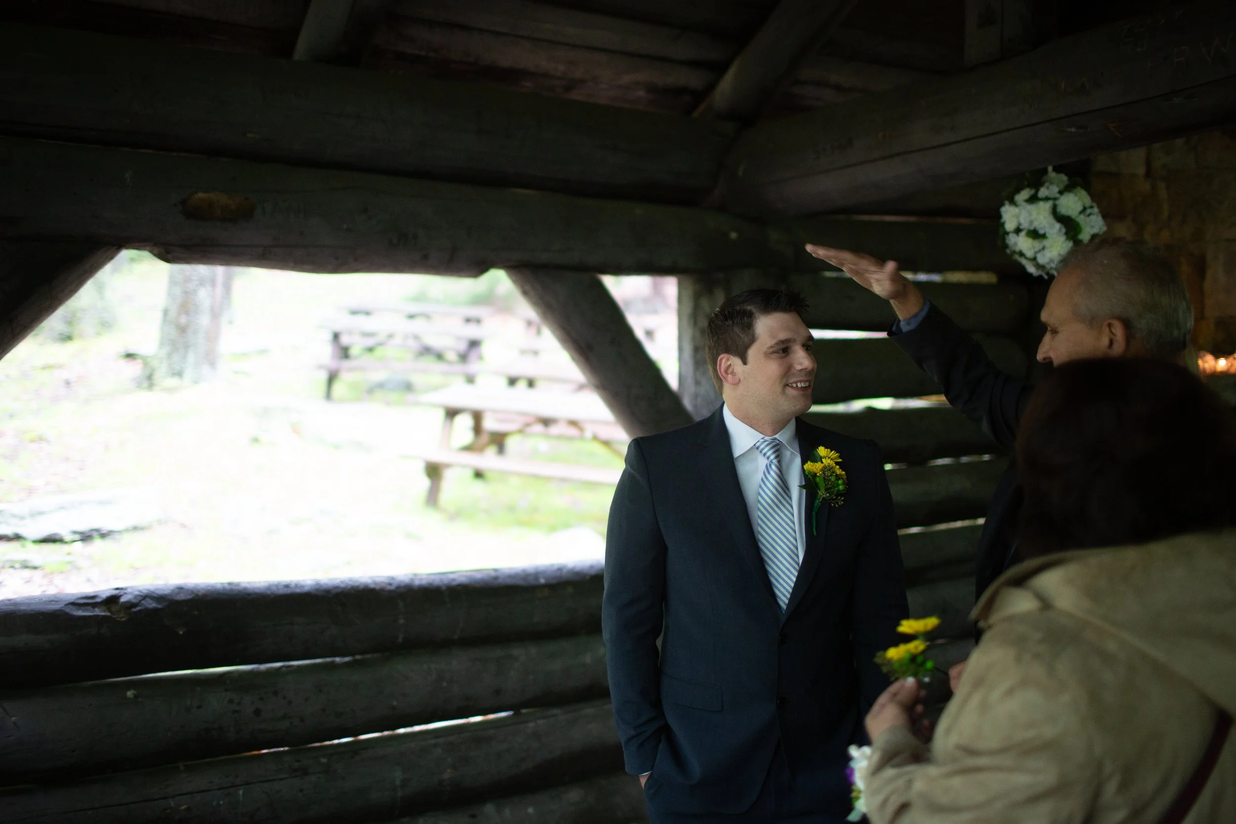 A groom in a suit smiling at an older woman, likely during a wedding ceremony, as she offers him flowers under a wooden shelter in an outdoor setting.