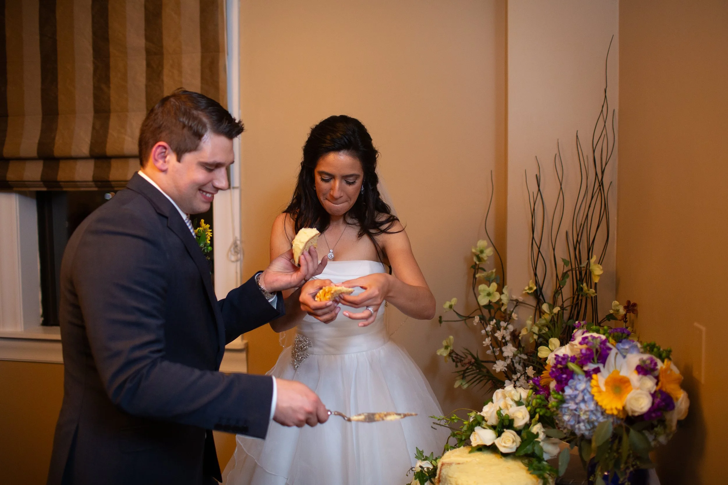 A bride and groom at a wedding reception, the groom holding a plate and the bride holding a piece of cake with her mouth, surrounded by flowers.