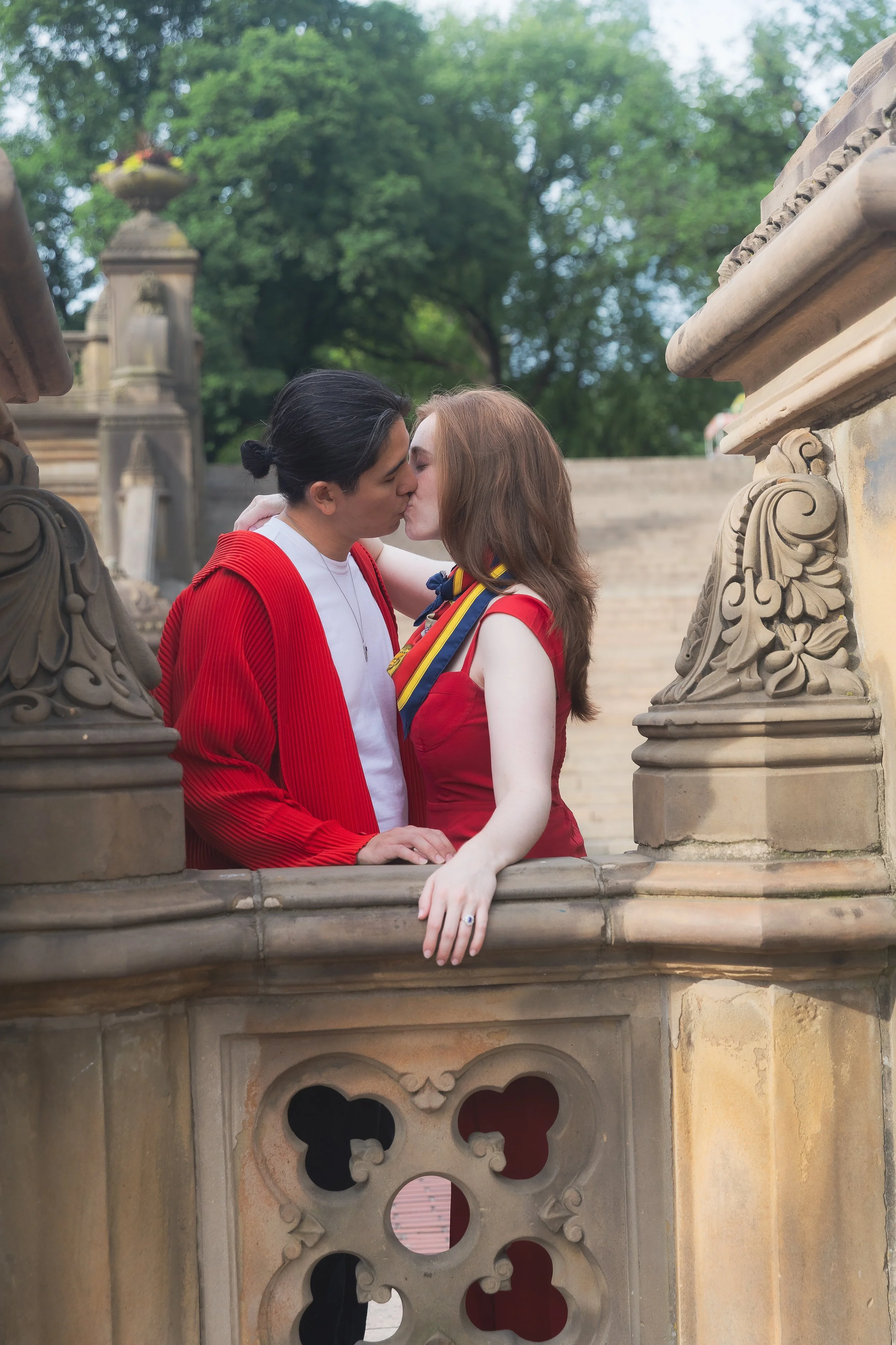A couple sharing a kiss on a stone balcony with carved decorative details, surrounded by lush green trees.