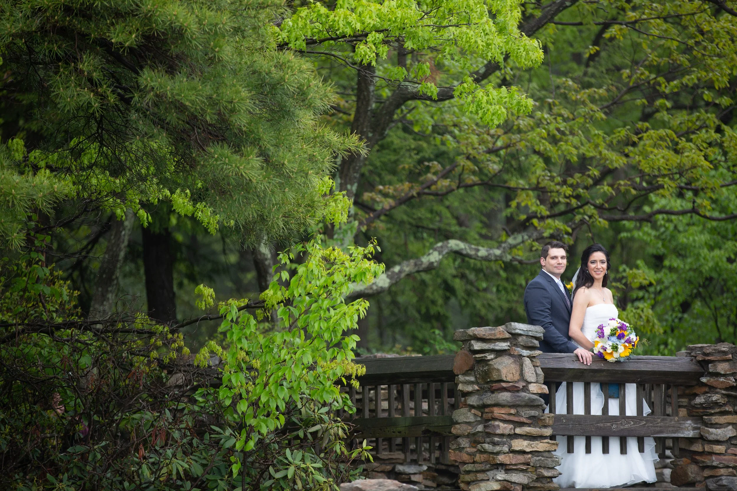 A couple dressed in wedding attire standing on a stone and wooden bridge in a lush green outdoor setting.