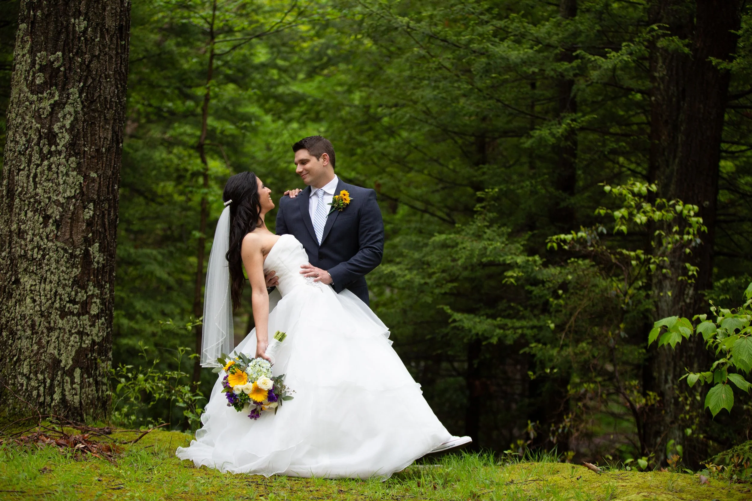 A bride and groom standing in a lush green forest, gazing at each other lovingly; the bride is holding a colorful bouquet, and the groom is wearing a dark suit with a sunflower boutonniere.