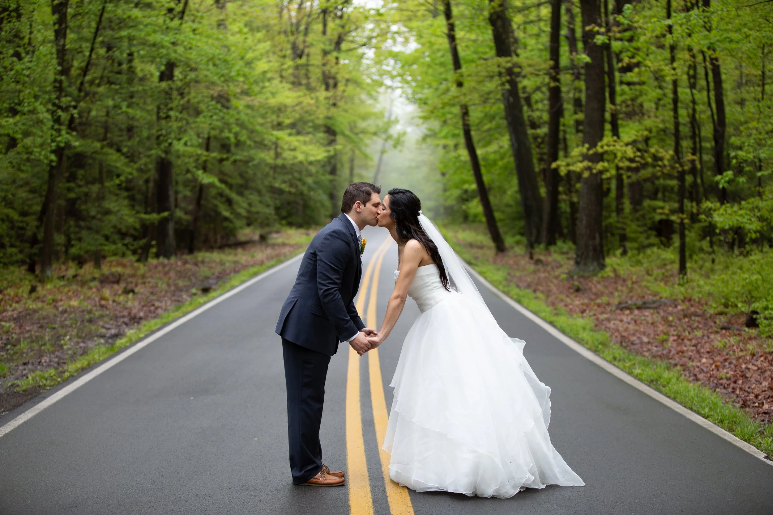 A bride and groom kissing and holding hands in the middle of a deserted forest road surrounded by lush green trees.