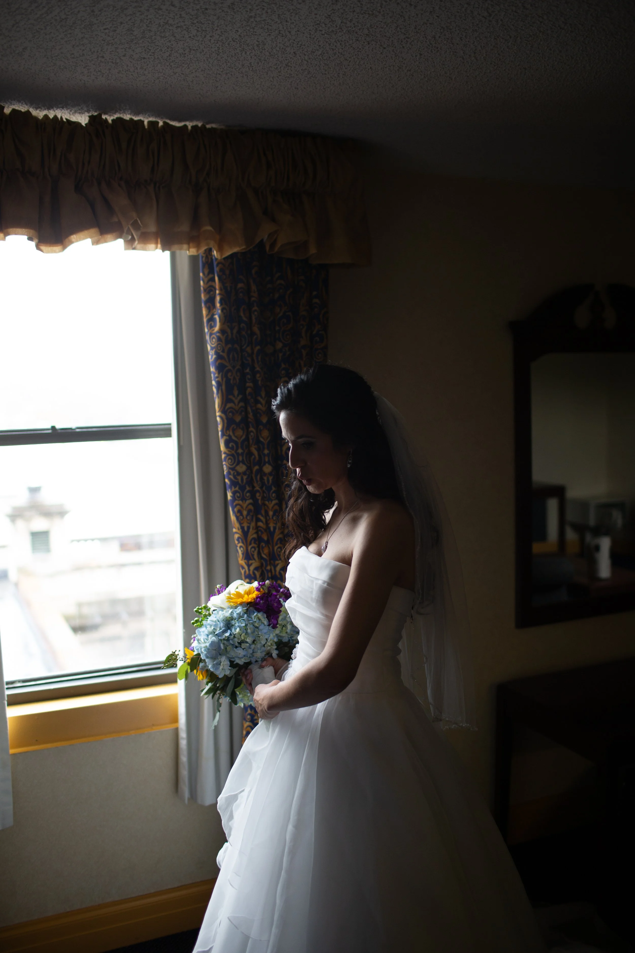 A woman in a white wedding dress holding a bouquet of flowers, standing by a window with patterned curtains, inside a room.