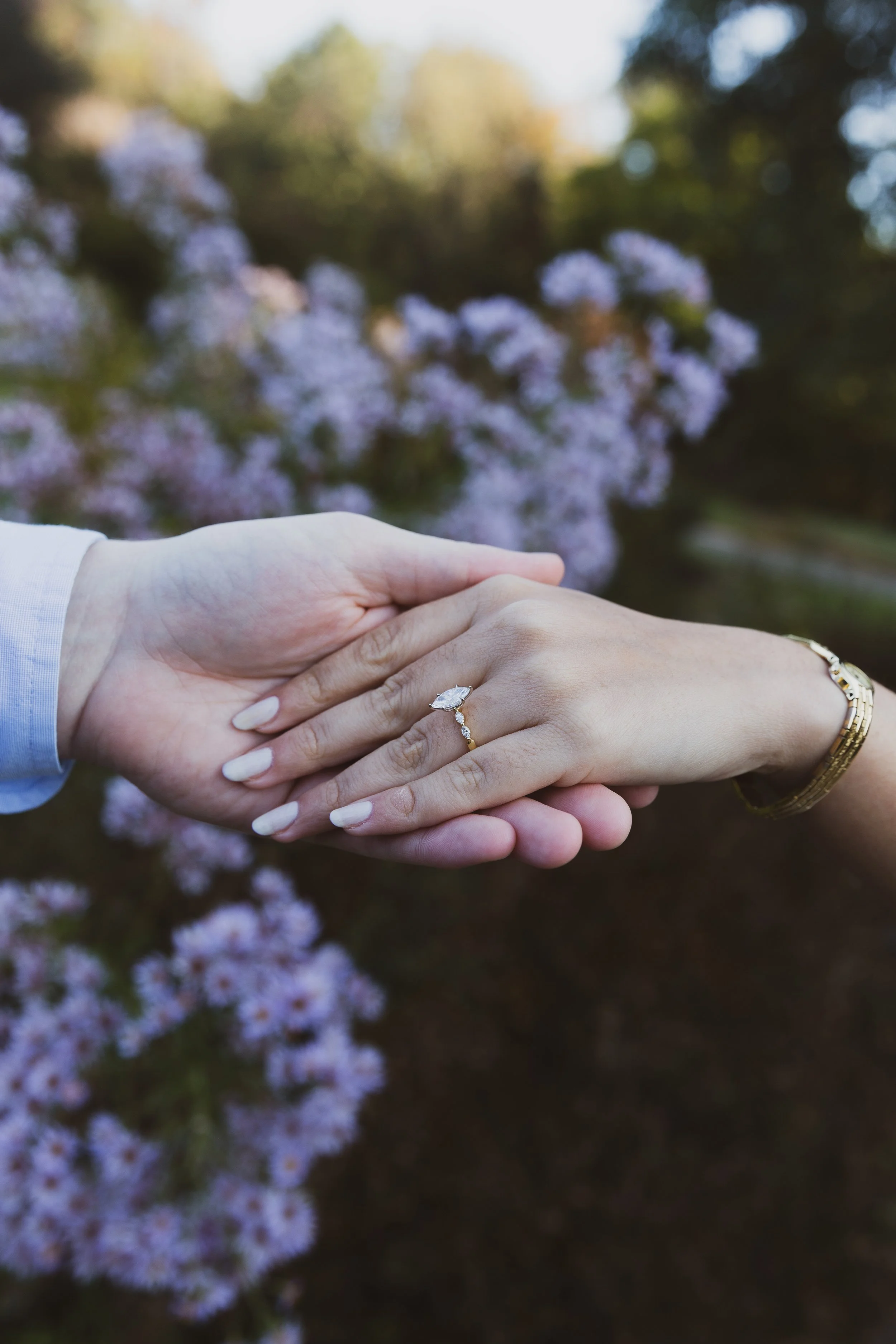 Two hands gently holding a woman's hand with a ring on her finger. One hand is wearing a gold bracelet. The background shows blurred purple flowers and green foliage.