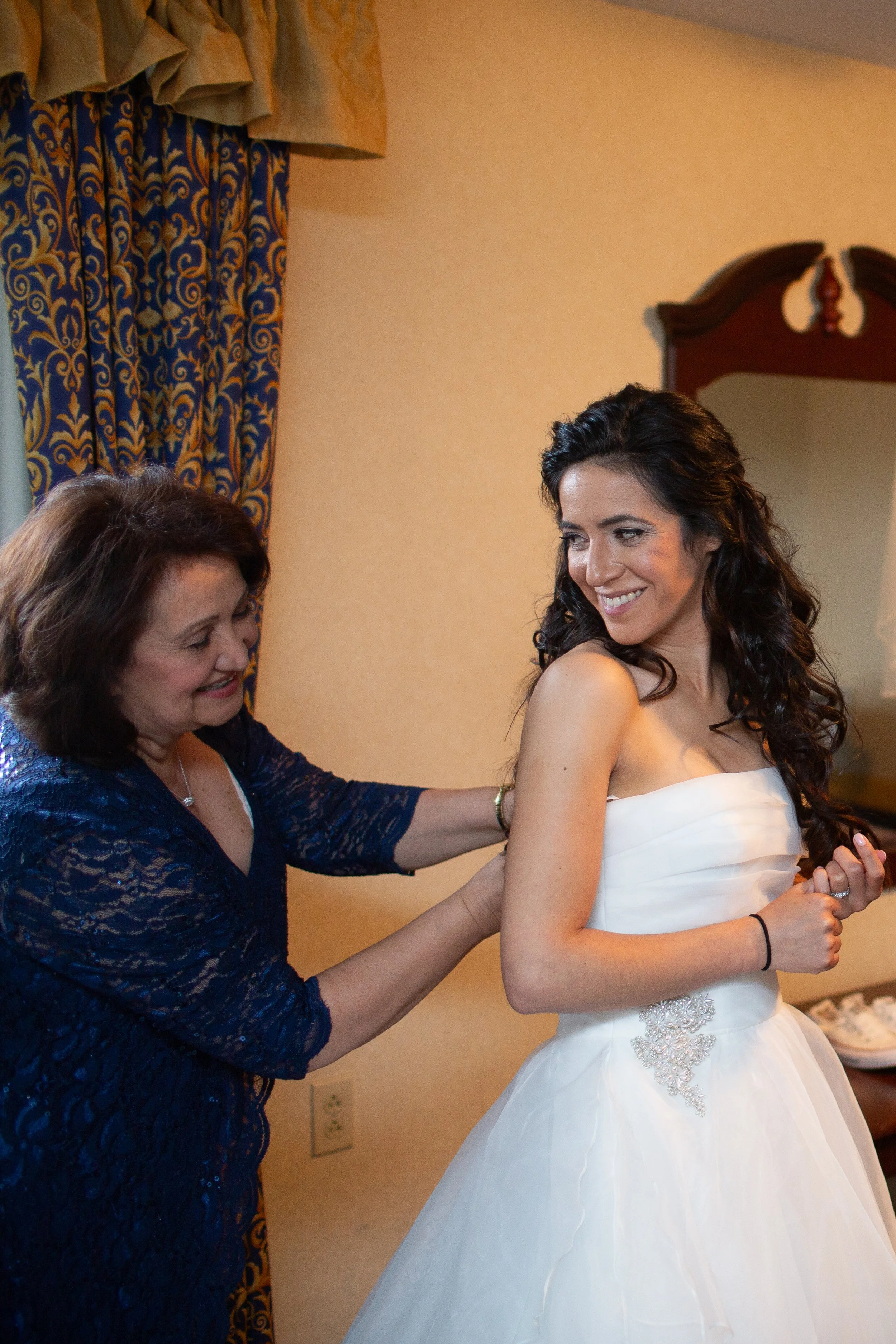 A woman in a wedding dress smiling as another woman helps her with her dress in a warmly lit room with blue and gold patterned curtains.