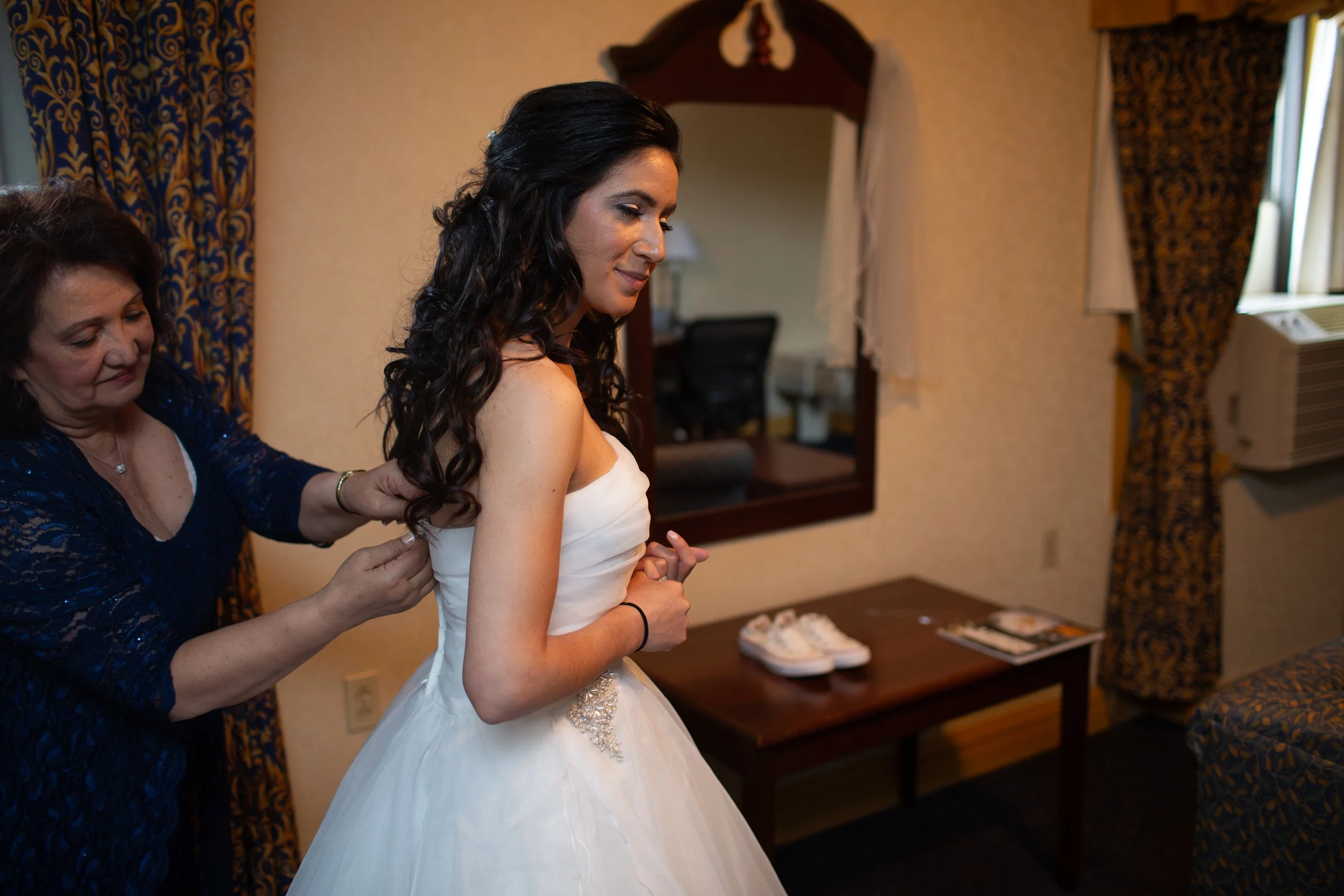 A bride in a white wedding dress is being helped by an older woman with dark hair in a blue dress, adjusting her dress. The bride has dark, curly hair and is smiling softly. The background shows a mirror and a table with some items, inside a room wit