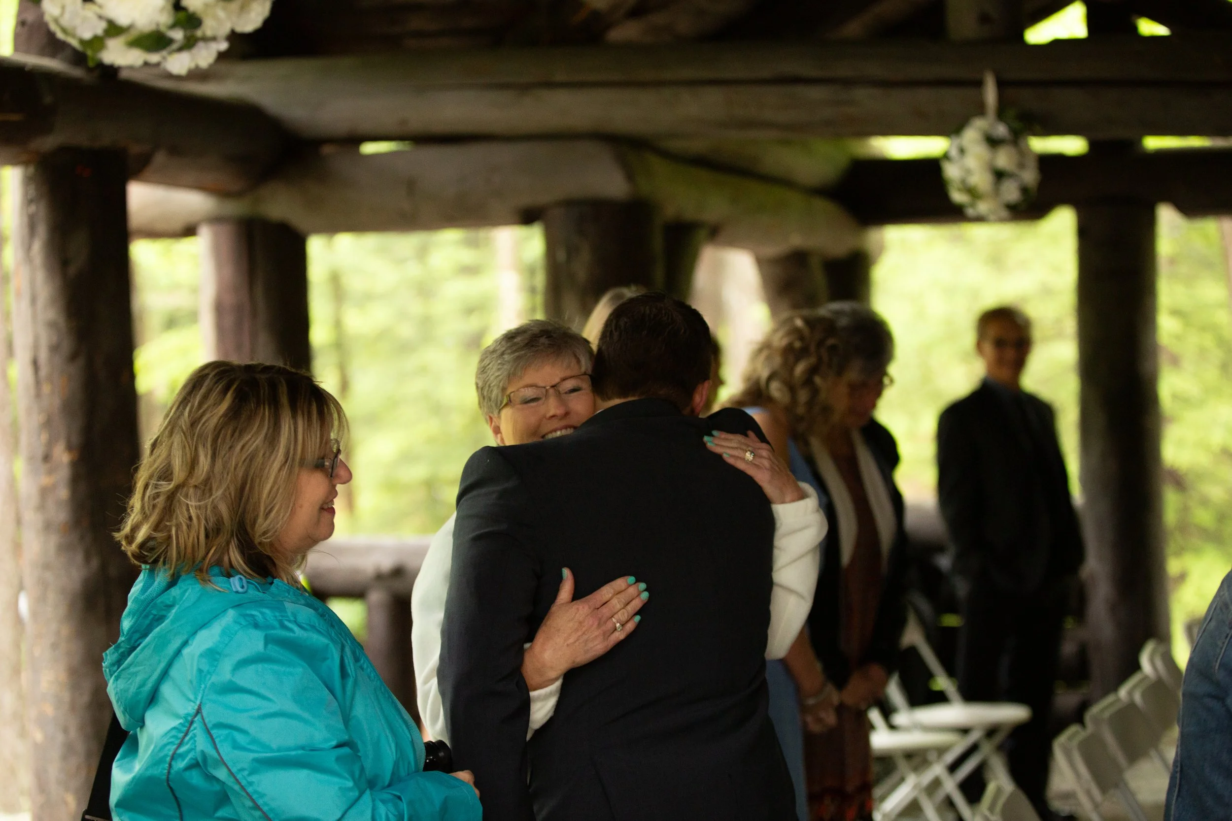 A woman in a blue jacket and glasses is praying and holding hands with a man in a dark suit under a wooden pavilion with green trees in the background.