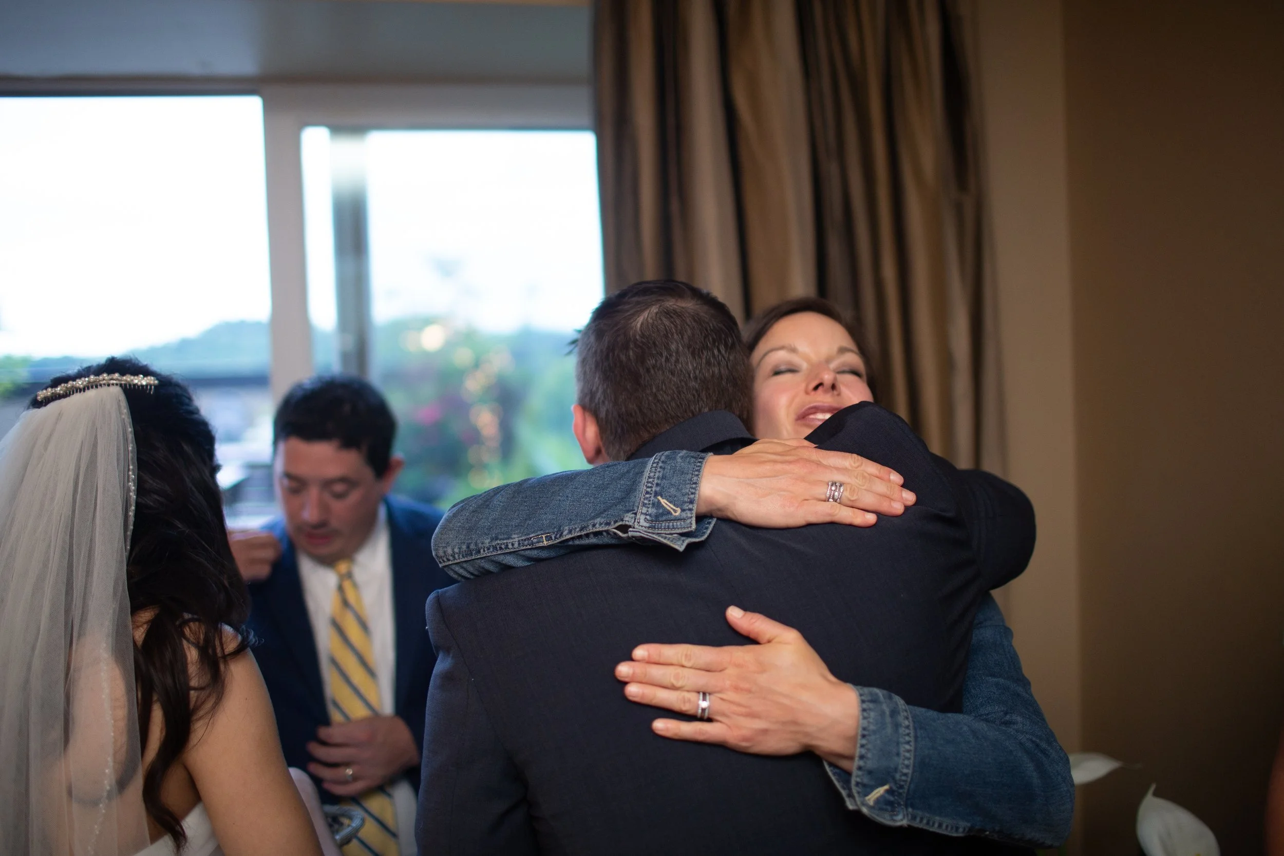 A happy couple hugging during a wedding ceremony inside a room with large windows and curtains.
