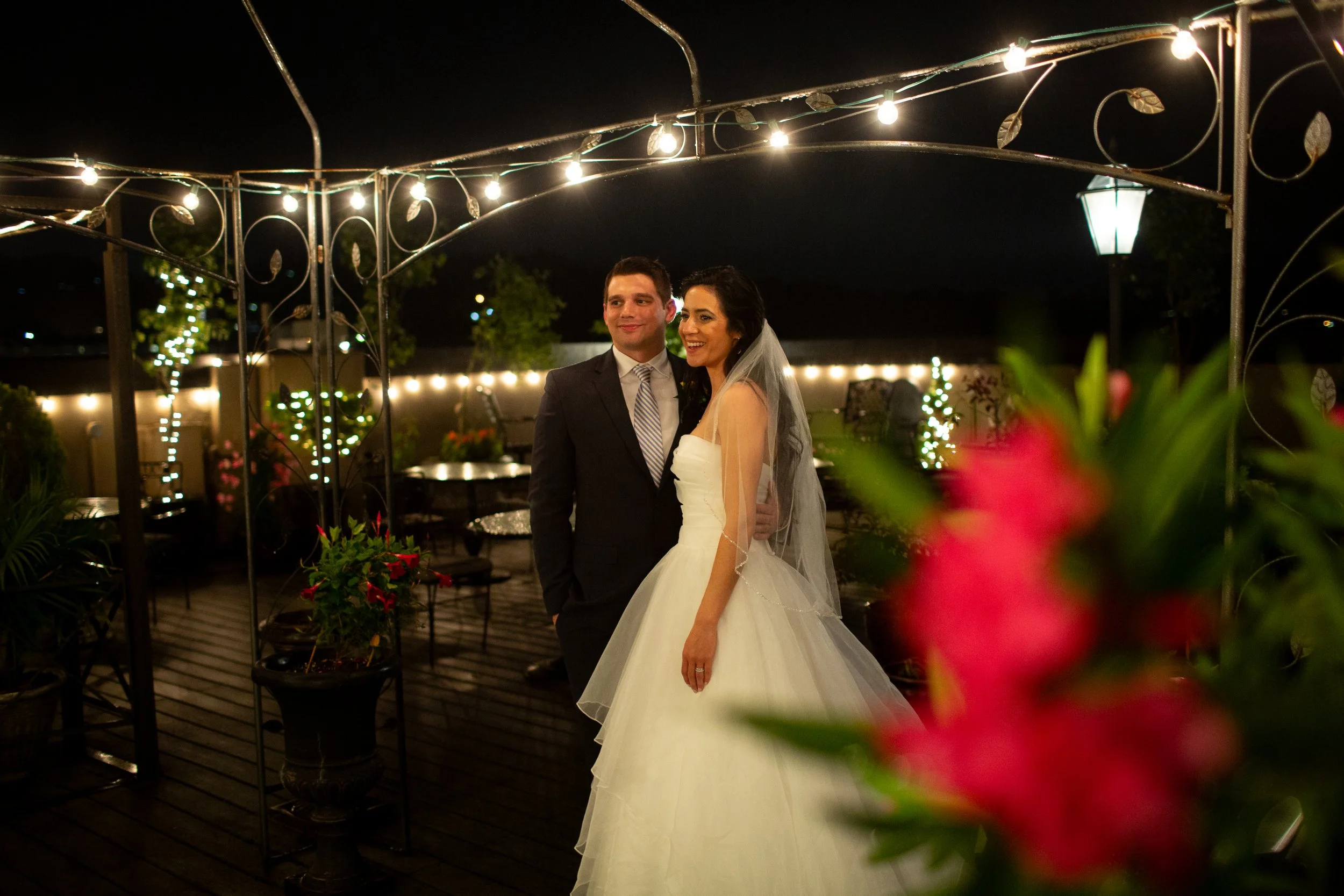 A bride and groom standing together on a decorated outdoor patio at night, illuminated by string lights and lanterns, with greenery and flowers around them.