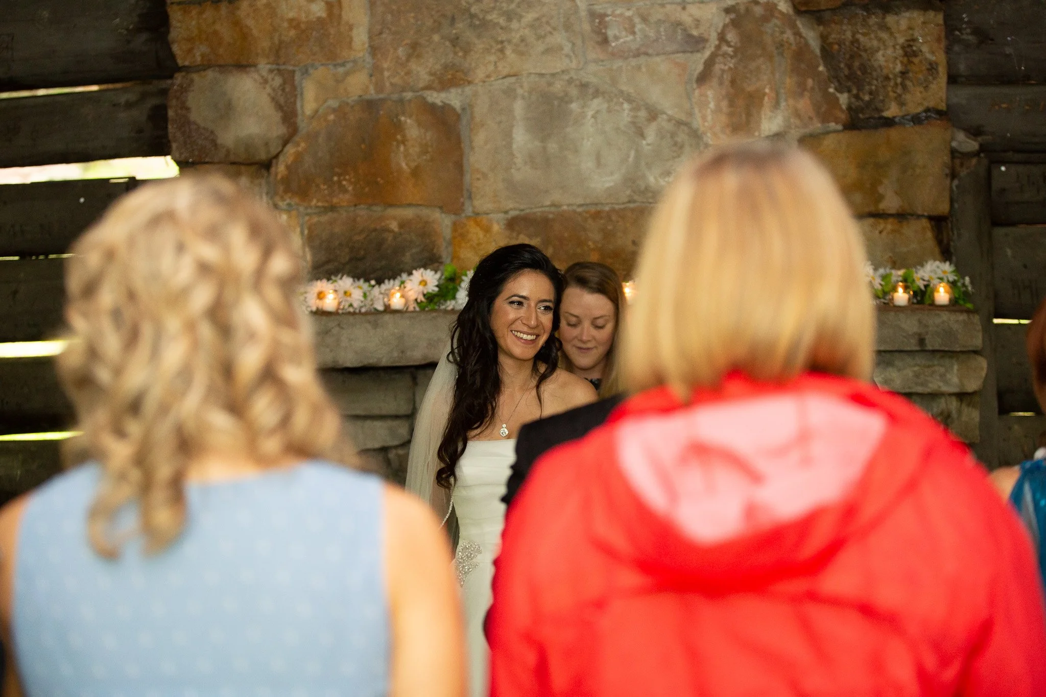 A woman in wedding dress smiling during a ceremony, surrounded by guests, with candles and flowers in the background inside a rustic stone wall venue.