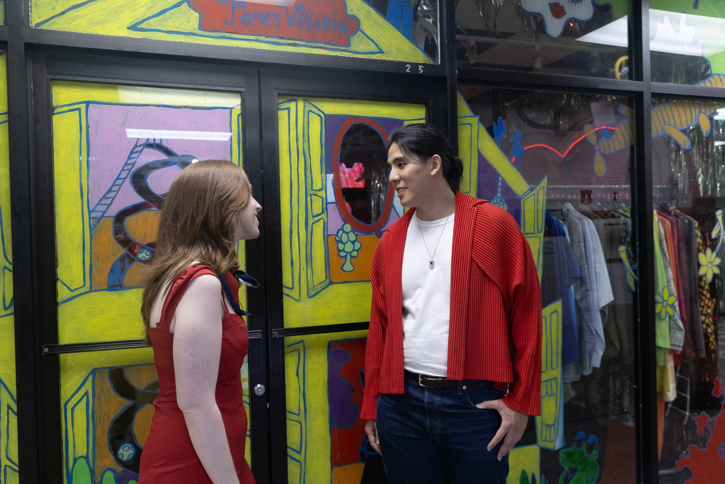 A couple conversing inside a store, with colorful artwork on the window behind them.