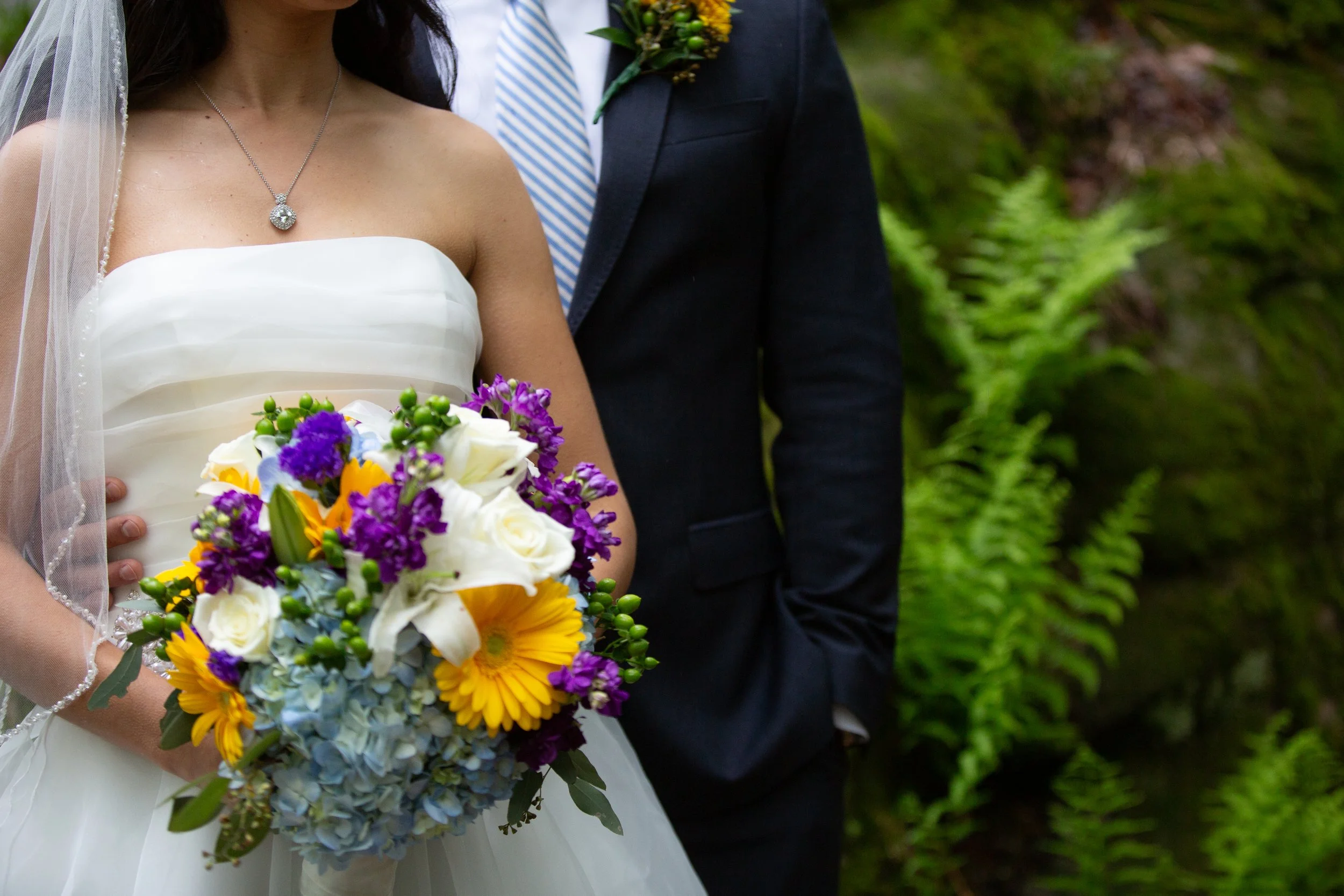 A bride holding a colorful bouquet of flowers with a groom standing beside her outdoors. The bride is wearing a white strapless dress with pleats and a diamond pendant necklace, and a veil. The groom is dressed in a dark suit with a white shirt, blue