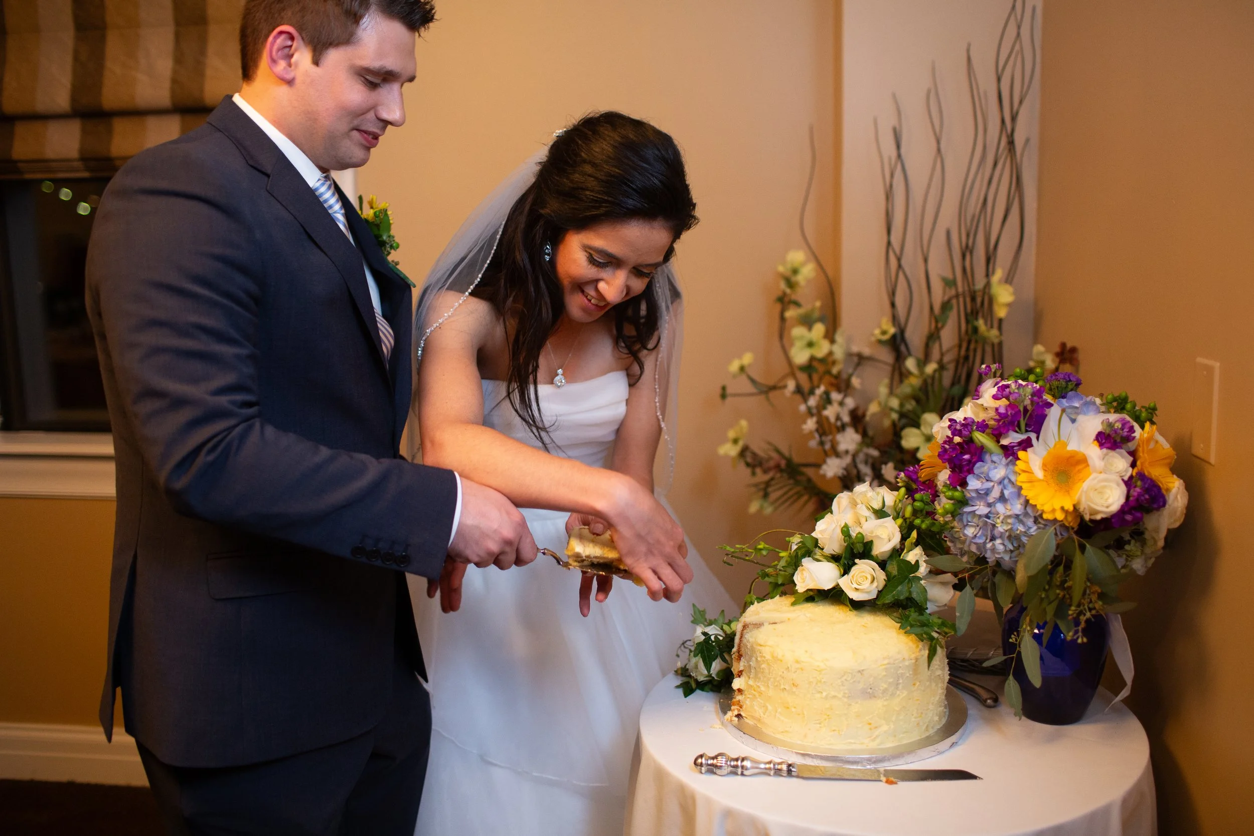 A bride and groom cutting their wedding cake, standing beside a table with a large floral arrangement.