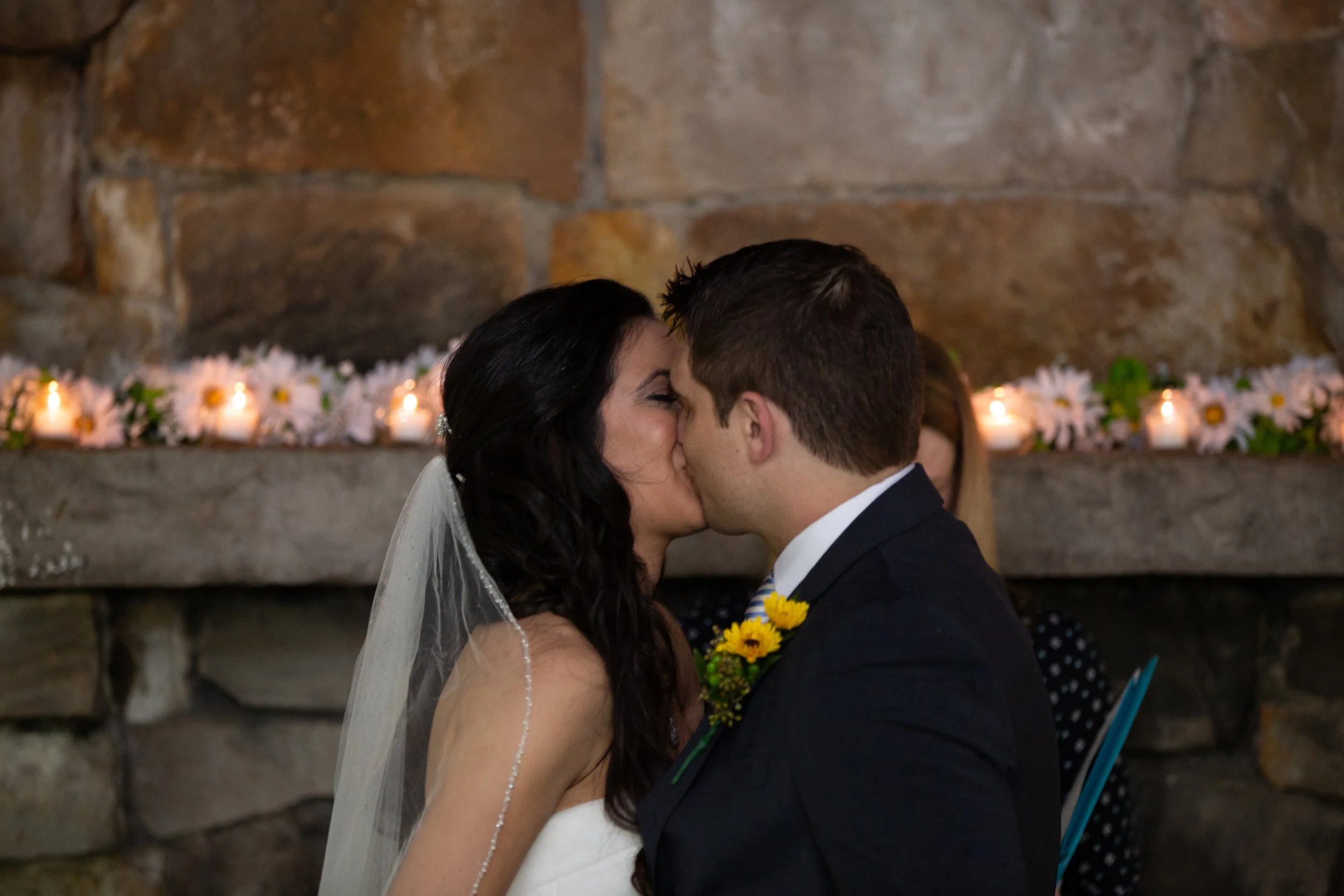 A couple kissing at their wedding ceremony in front of a stone fireplace decorated with candles and flowers.